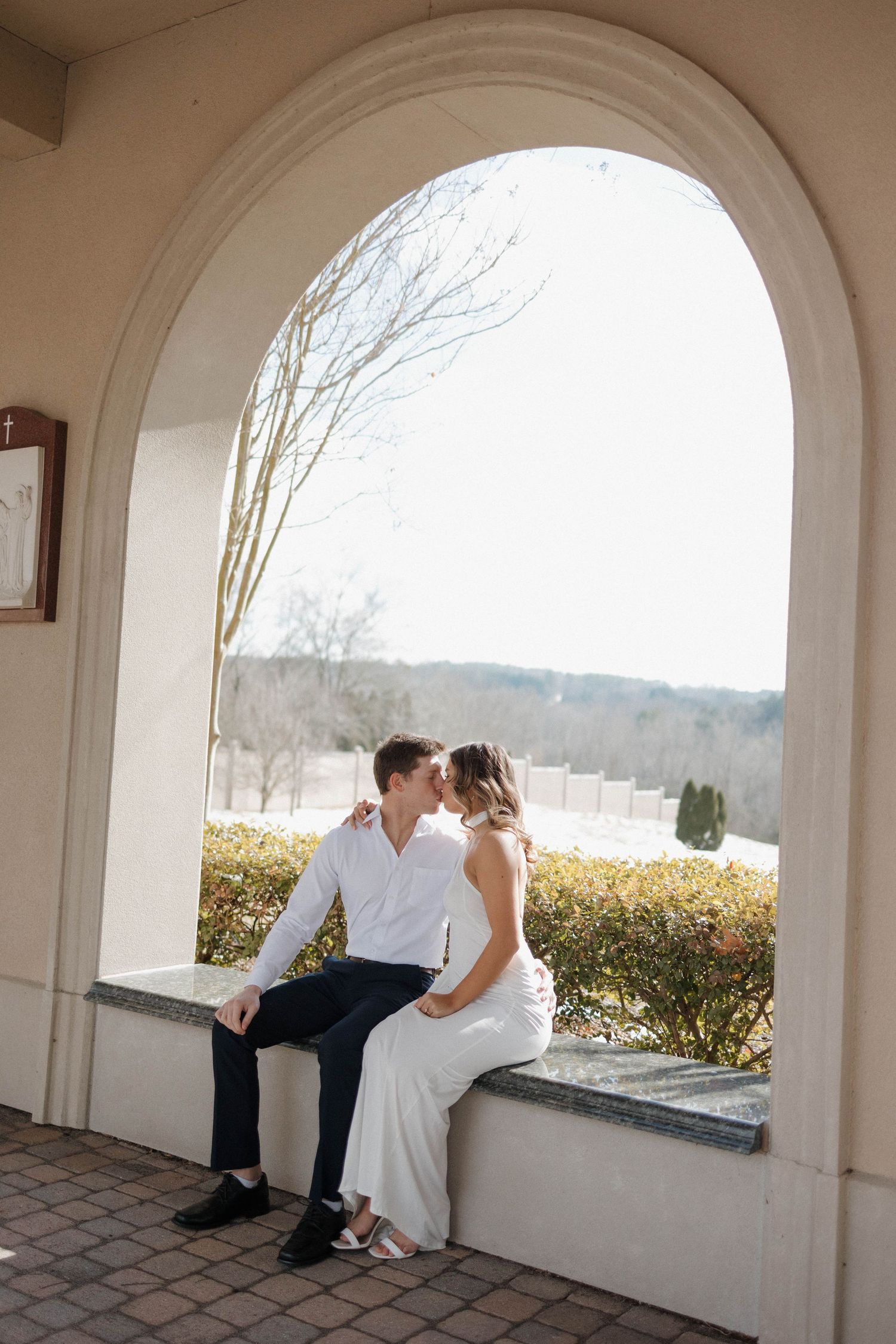 Elegant couple in formal wear sits together on a stone ledge beneath white arches.