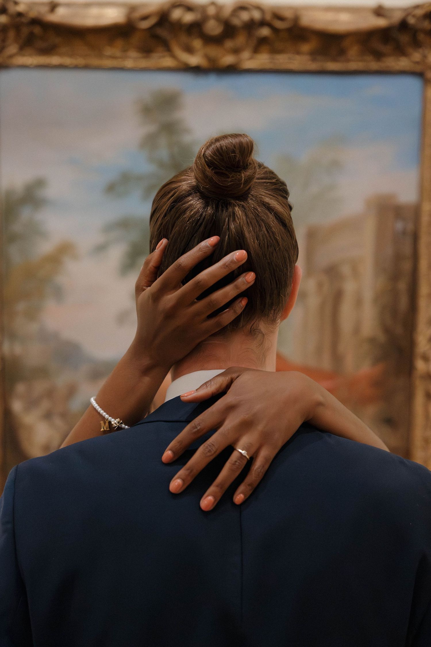 Intimate embrace showing hands wrapped around neck against classical painting backdrop.