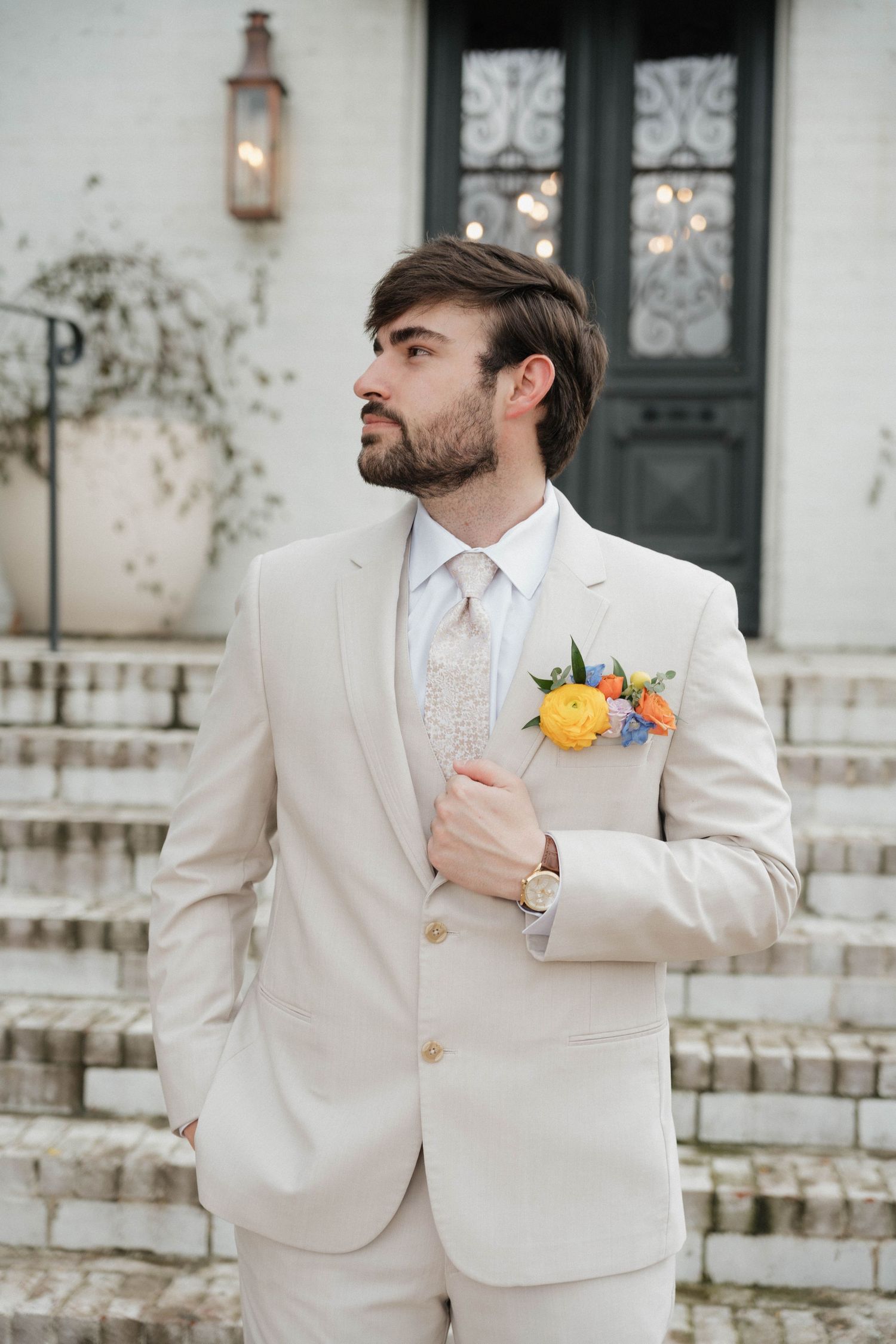 Groomsmen in light beige suits with colorful boutonnieres stand by ornate doorway.
