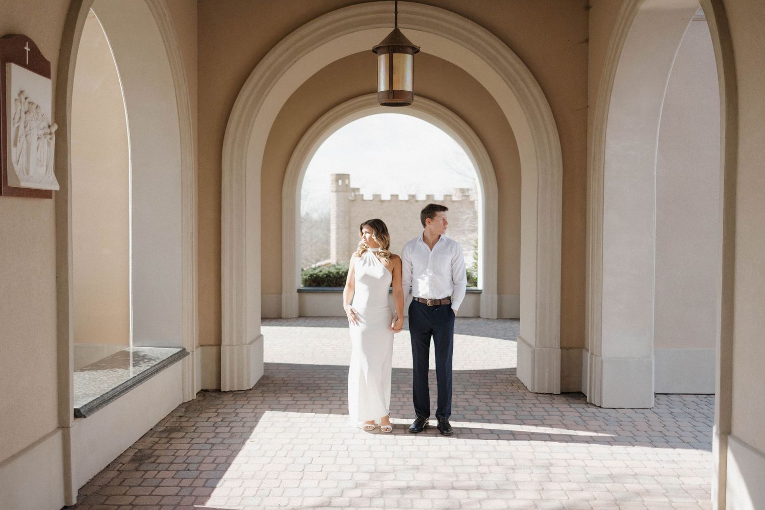 Elegant couple stands in sunlit arched hallway with castle visible in background.