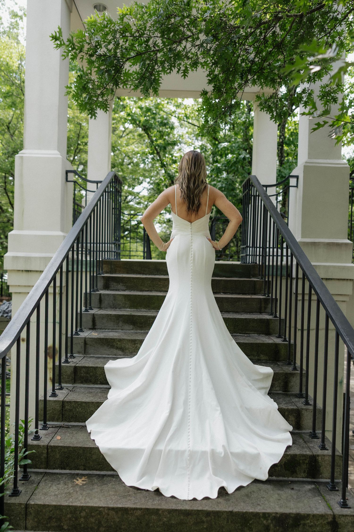 Bride twirling in flowing white gown on stone staircase surrounded by columns and greenery.