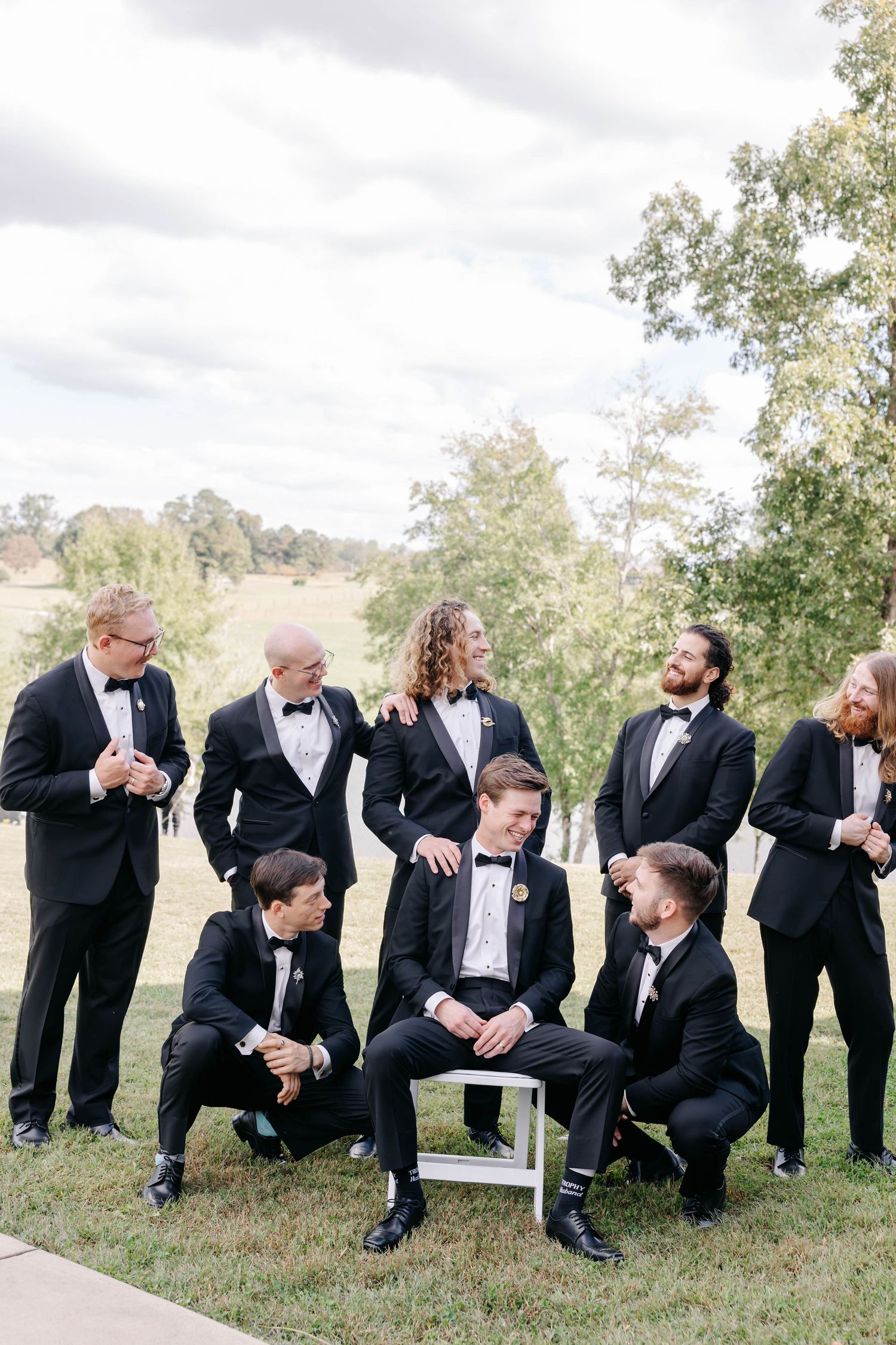 Groomsmen in black tuxedos gather casually on grassy hillside for group photo.