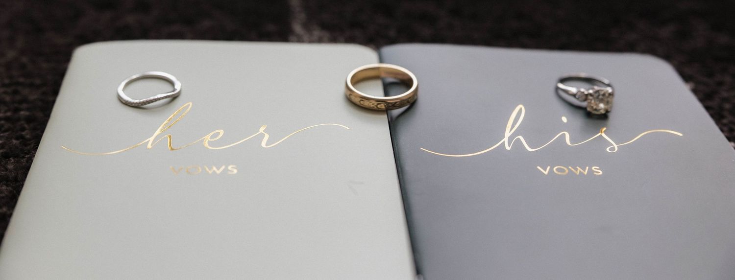 Two wedding rings and an engagement ring resting on matching gray her vows and his vows books.