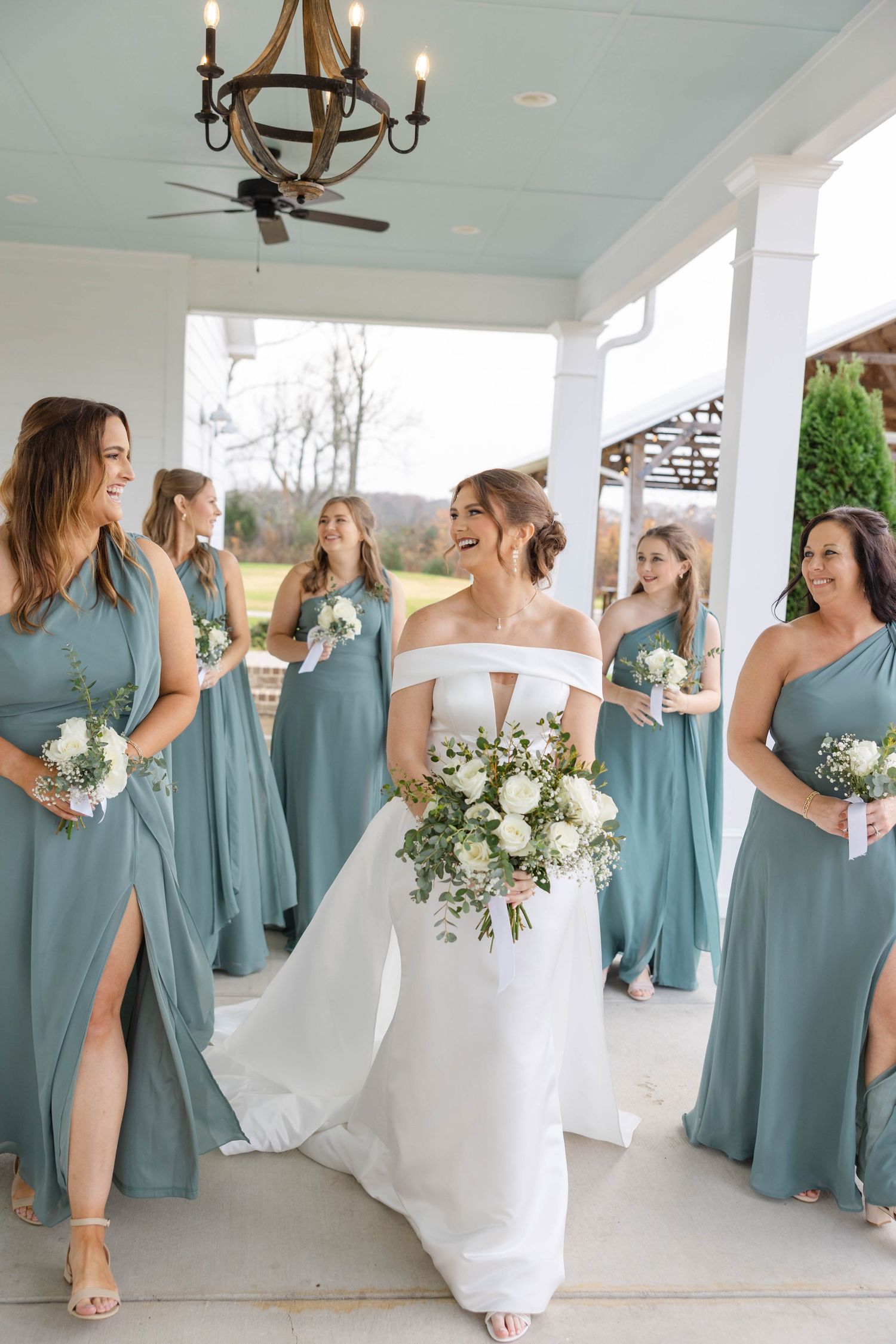 Bridesmaids in dusty blue dresses laughing with bride in white gown on covered porch.
