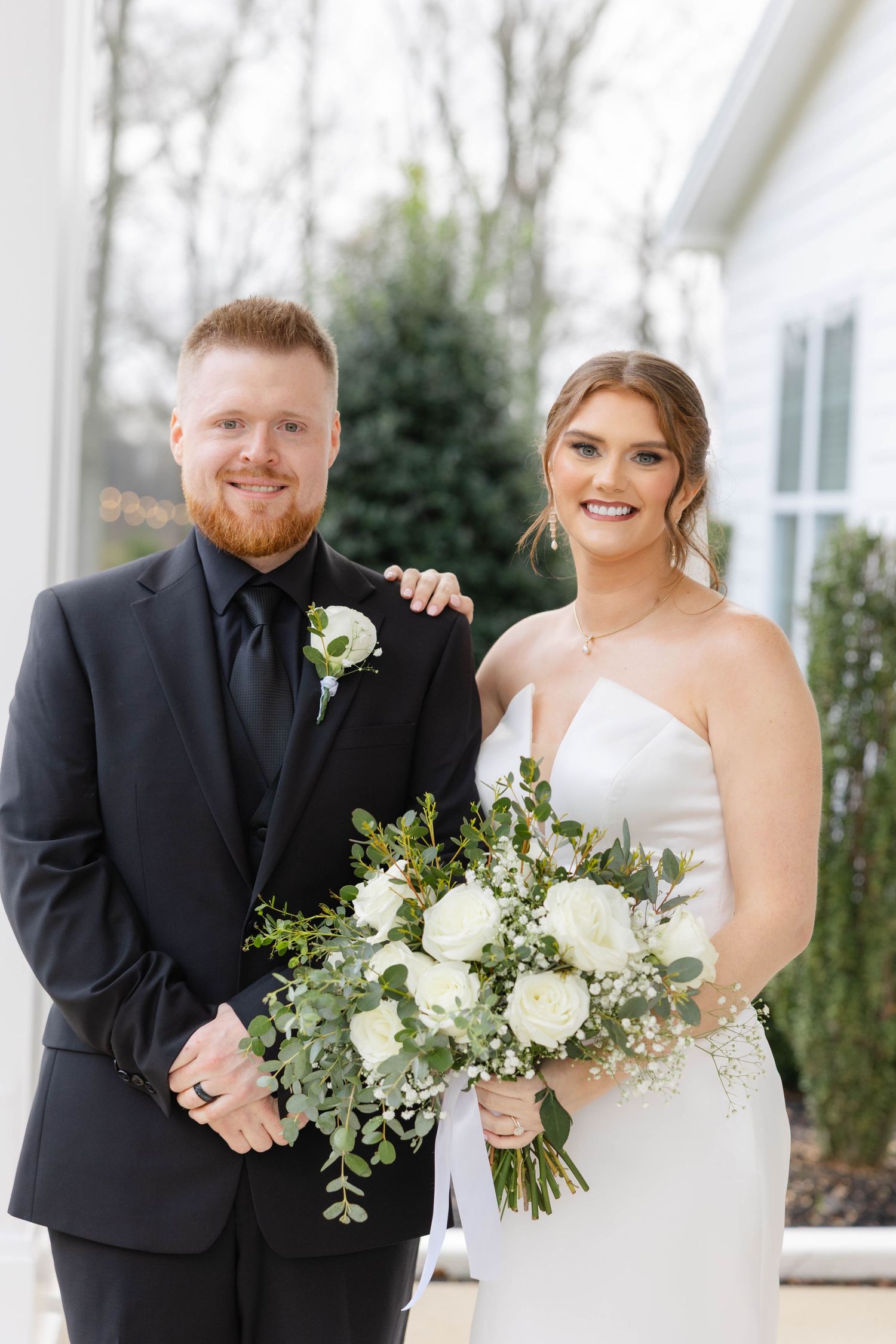 Wedding couple posing with white floral bouquet on covered porch.
