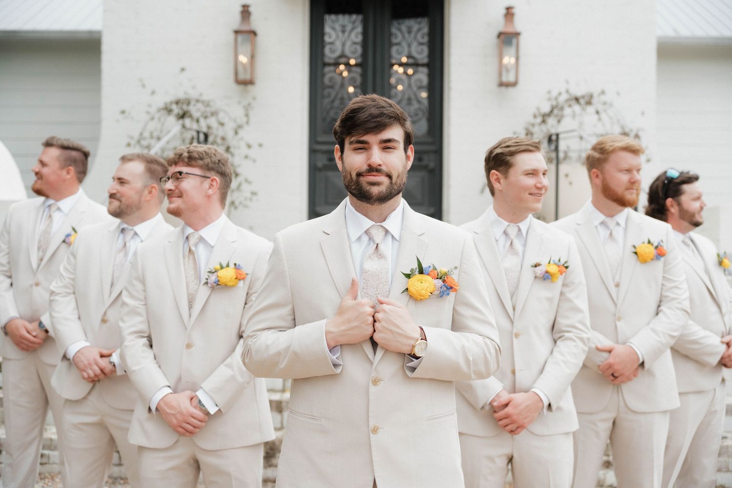Groomsmen in cream suits line up against white building exterior.