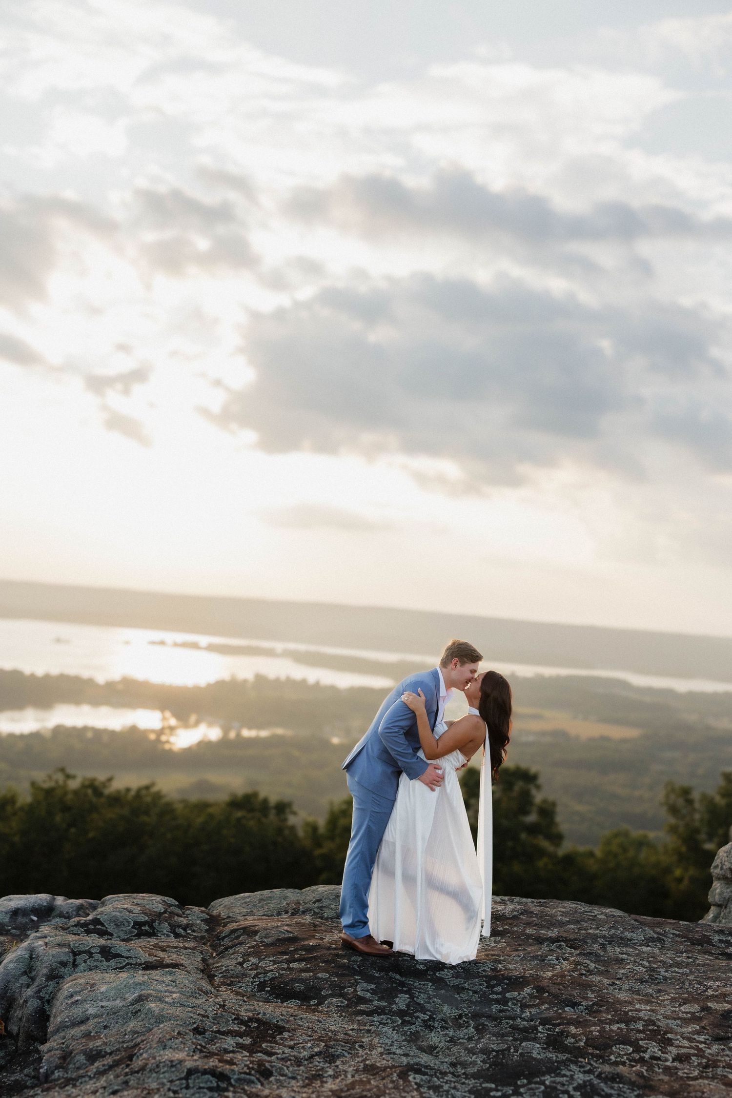 Couple shares a romantic kiss on a rocky cliff overlooking a scenic valley at dusk.