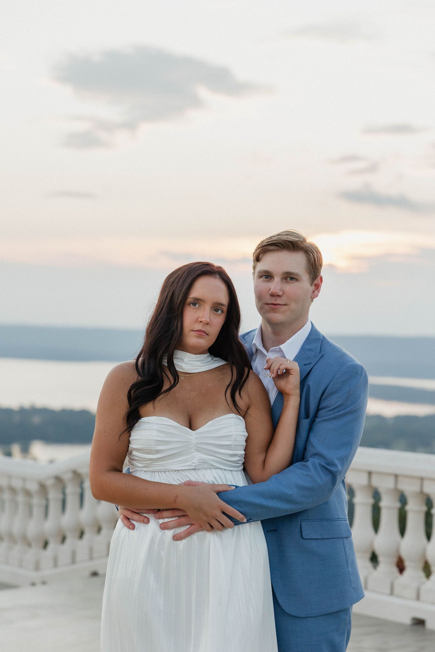 Couple in formal attire posing on white balcony during sunset.