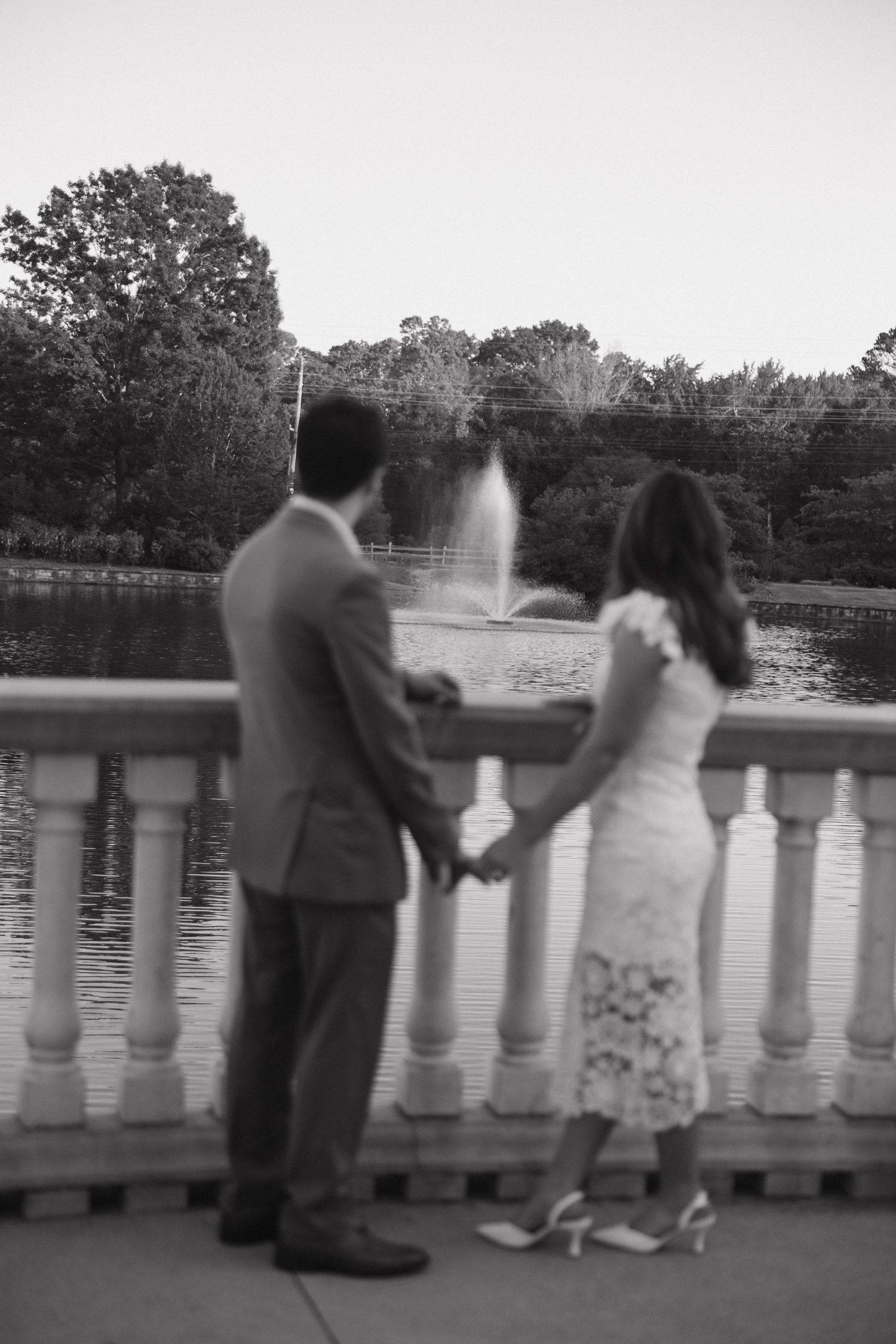 Black and white photo of a couple holding hands on a bridge overlooking a fountain and lake.