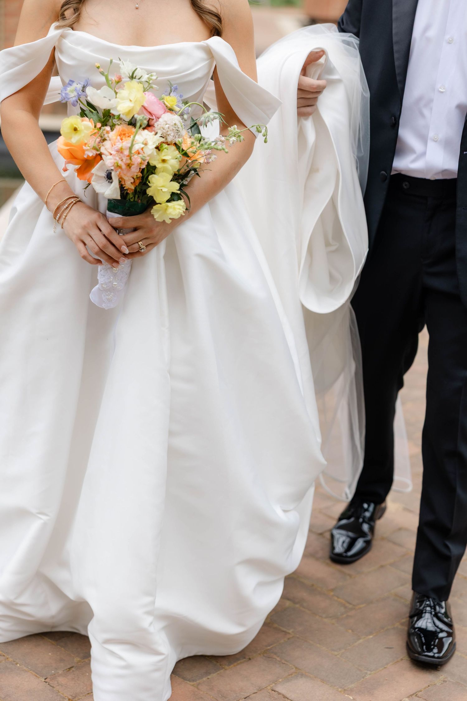 Bride in white off-shoulder ball gown holding colorful spring bouquet while groom in black tuxedo holds her train.