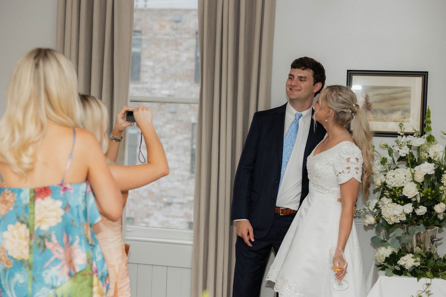 Wedding guests mingle and celebrate in a reception room decorated with floral arrangements.