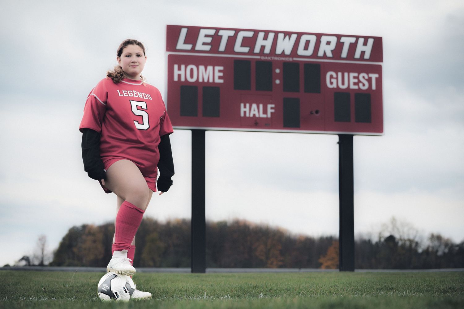 Soccer player in red uniform rests foot on ball beneath scoreboard at Letchworth field.