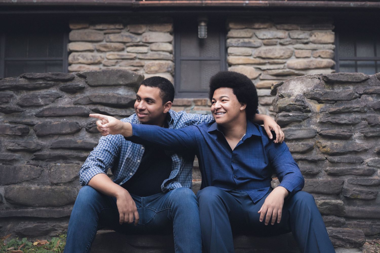 Two friends in denim outfits sit on stone steps while sharing a lighthearted moment together.