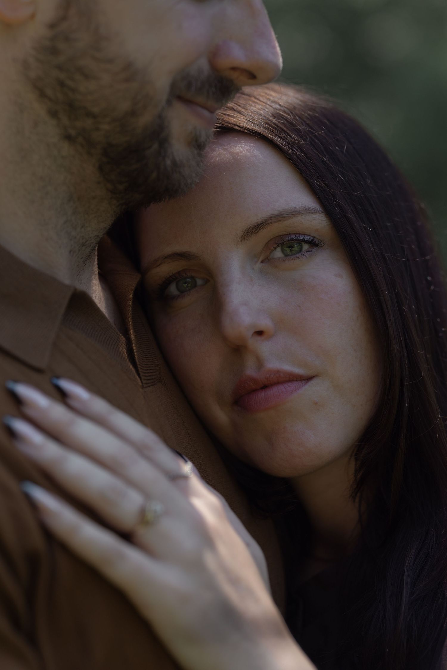 A woman with dark hair rests her head on a man's chest, showing an engagement ring, in a romantic outdoor portrait.
