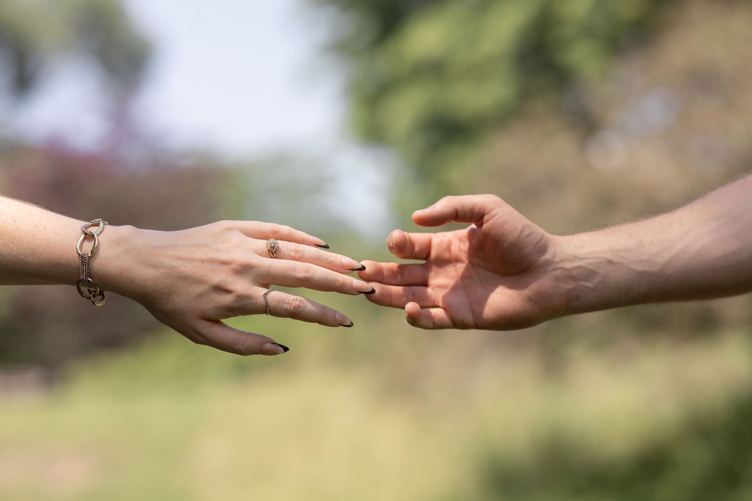 Two hands reaching toward each other outdoors, one with a bracelet and ring, against a green blurred background.