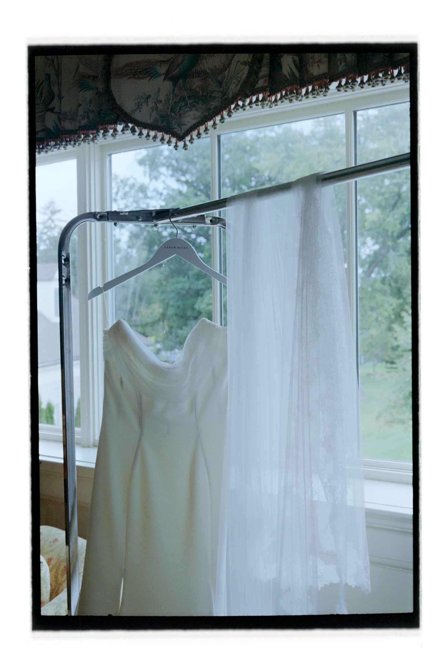 White wedding dress hangs by window with sheer curtains.  Rochester NY Film wedding photography