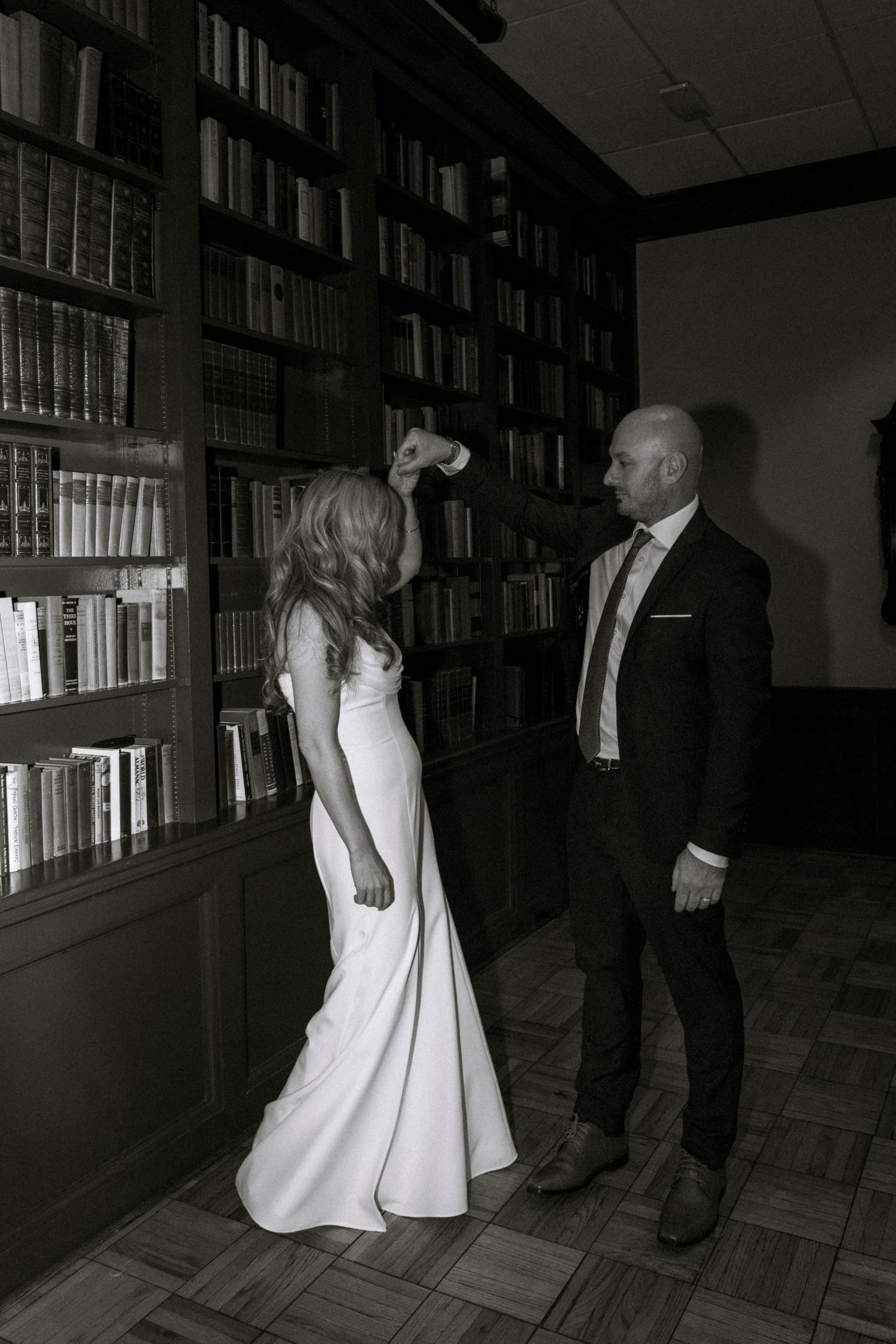 A couple poses in formal attire against a wall of bookshelves in dramatic lighting.
