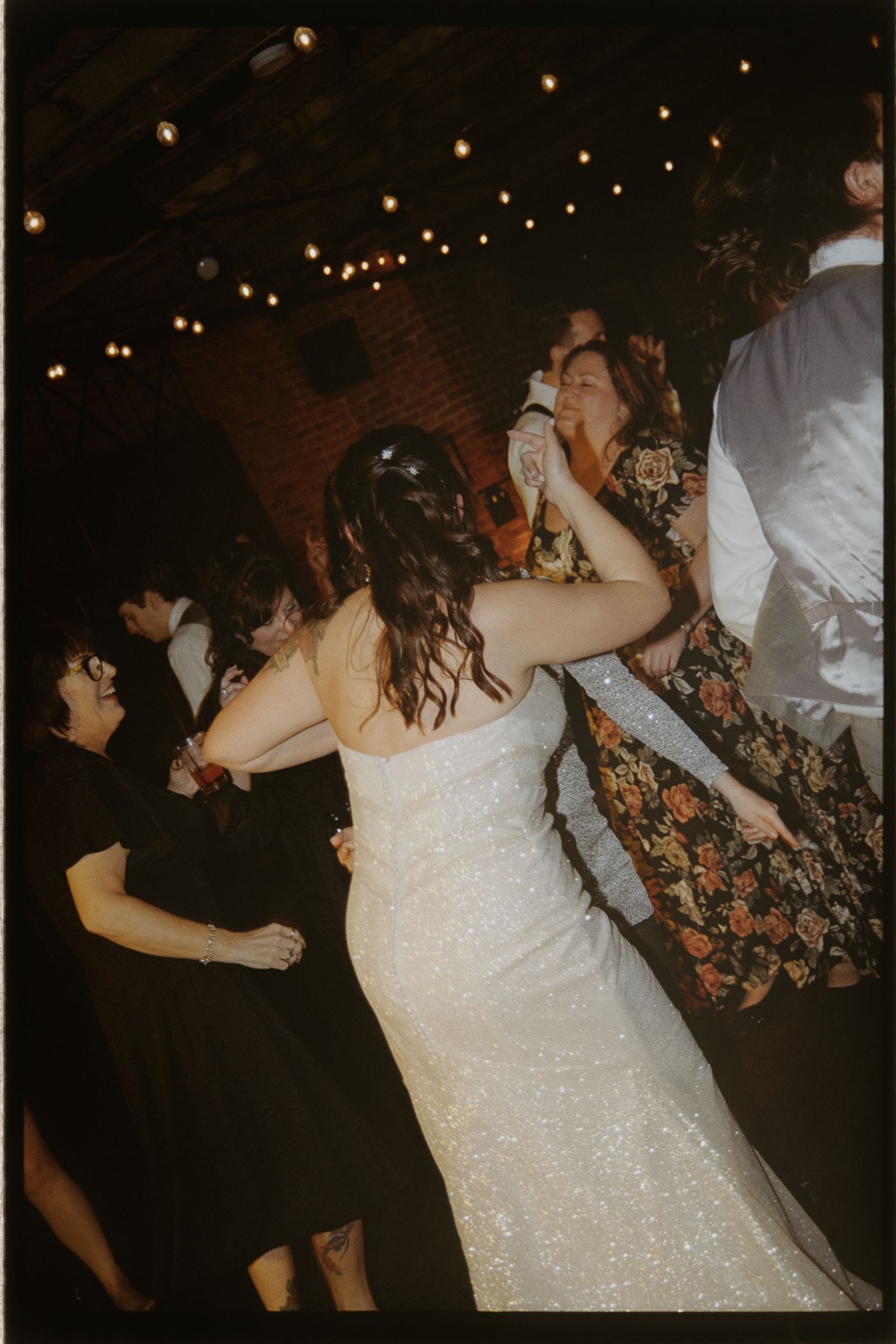 People dancing and celebrating at a wedding reception under twinkling lights with the bride in a sparkling white dress.