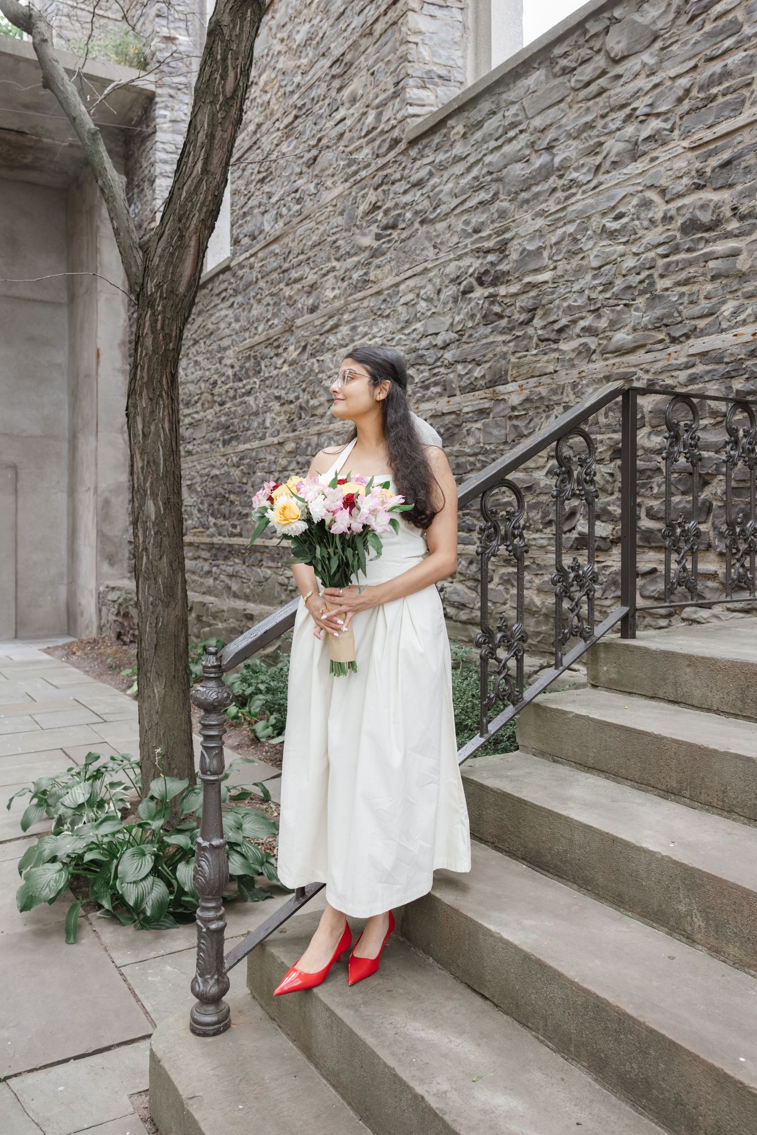 A woman in a white dress stands on stone steps, holding a bouquet of flowers, in front of a stone wall.