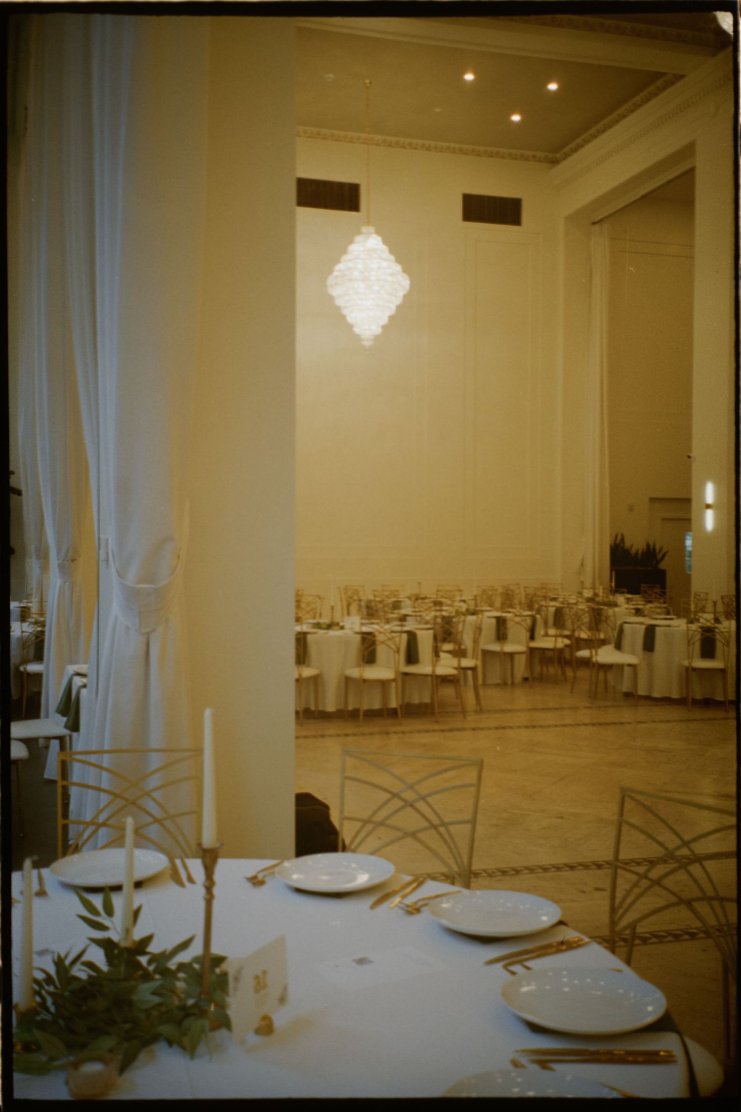 Empty wedding reception tables set with white linens and centerpieces under a crystal chandelier. Rochester NY film wedding photographer