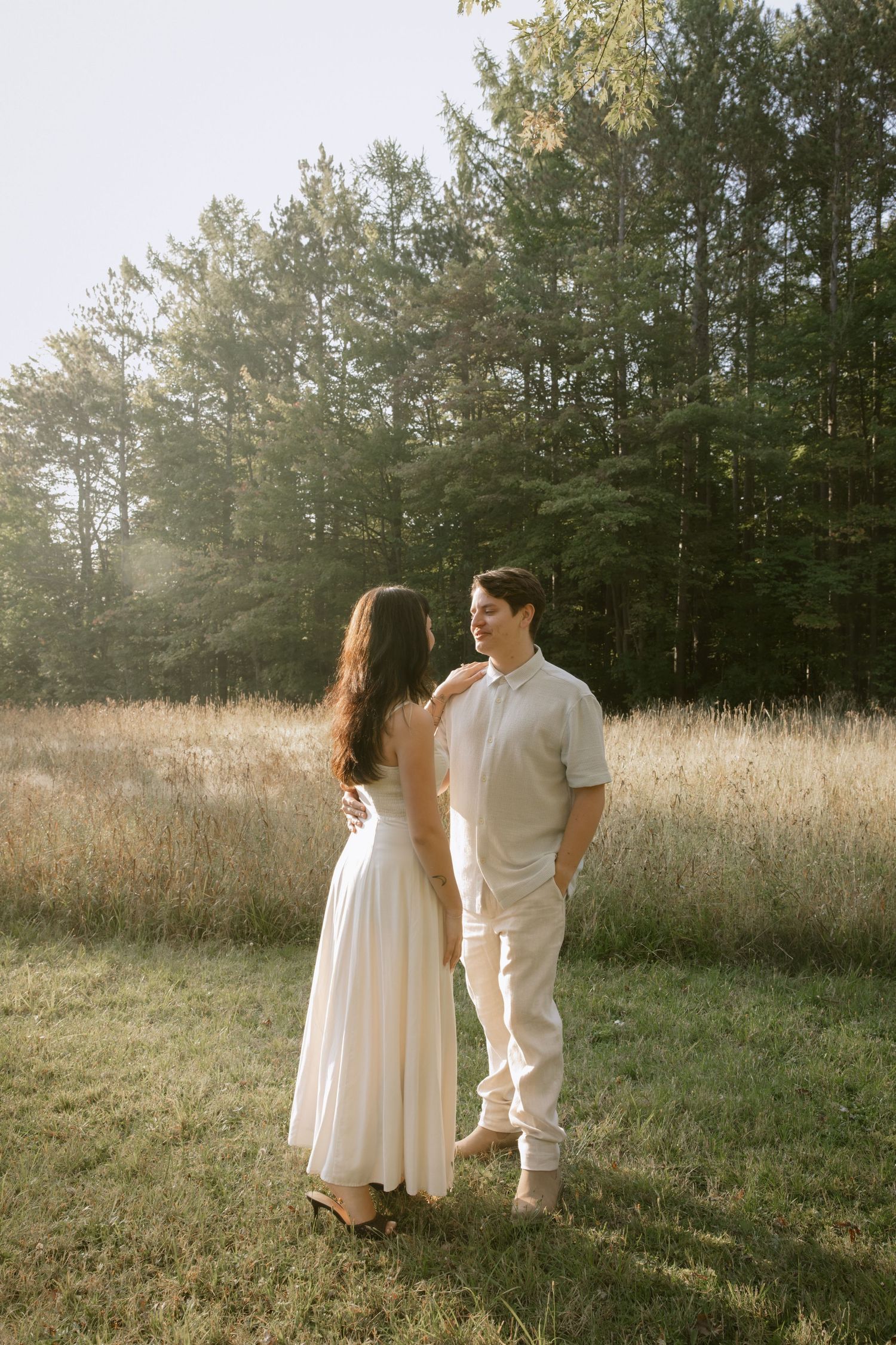 A couple in white outfits stands together in a sunlit meadow with tall grass and pine trees in the background.