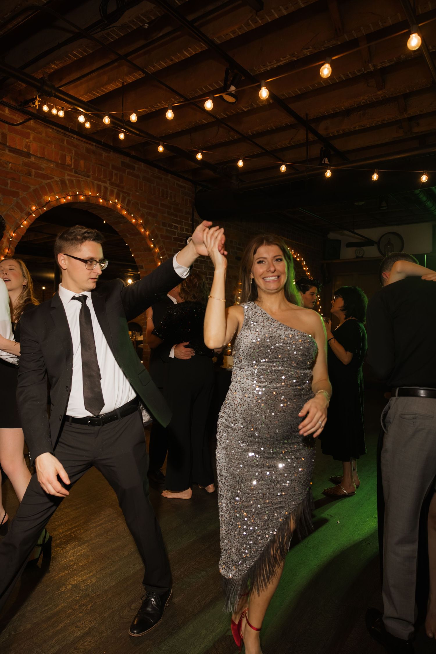 A well-dressed couple dancing at a formal event, with the woman wearing a sparkling silver dress and the man in a suit.