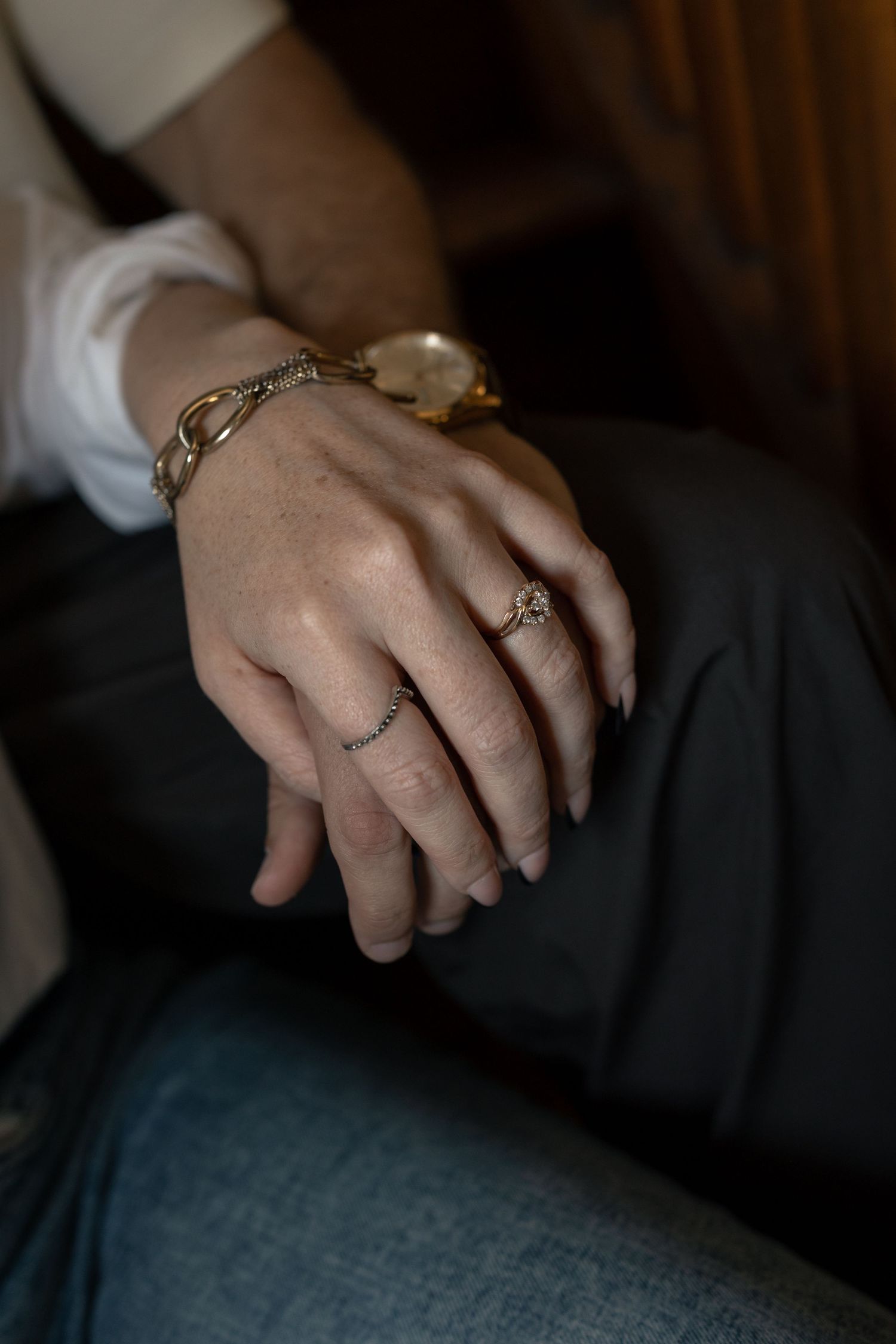 Close-up of woman's hands adorned with gold chain bracelet, vintage watch, and diamond ring resting on dark pants.