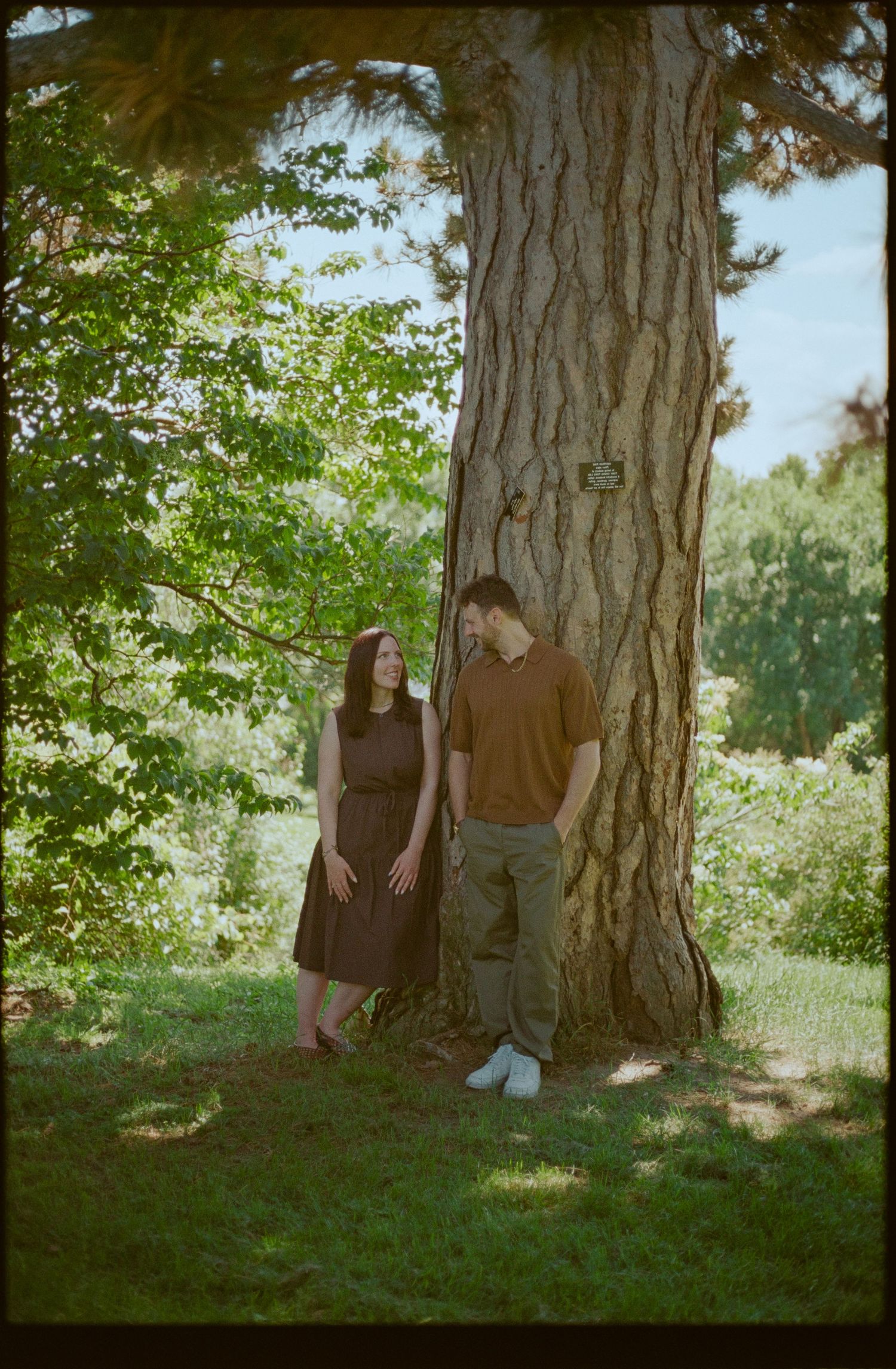 Couple leaning against a large tree trunk in a lush green park, smiling at each other on a sunny day.