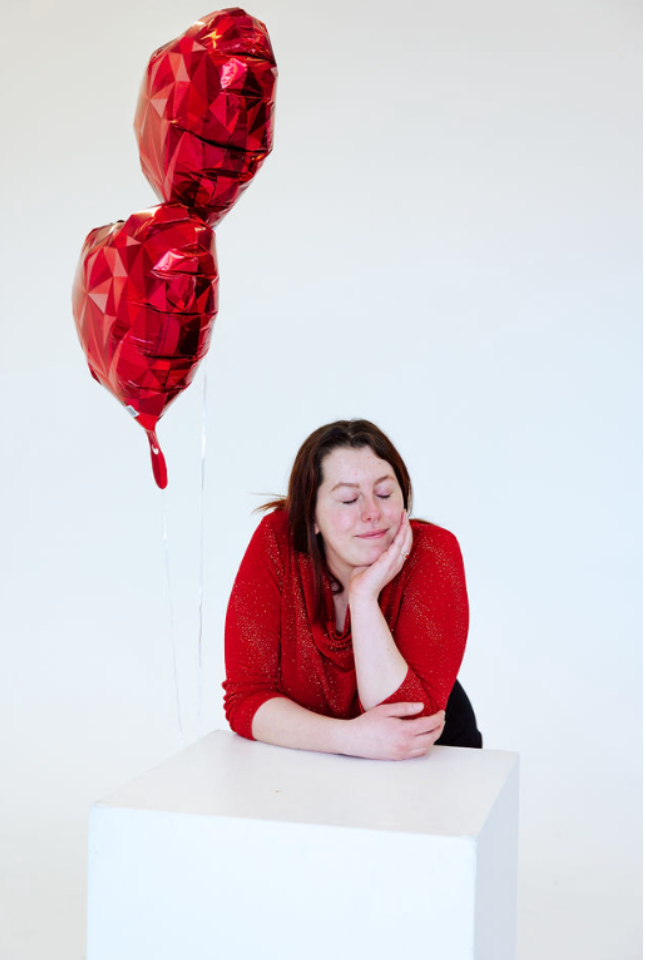 Person in red sweater posing with heart-shaped balloons against white background.