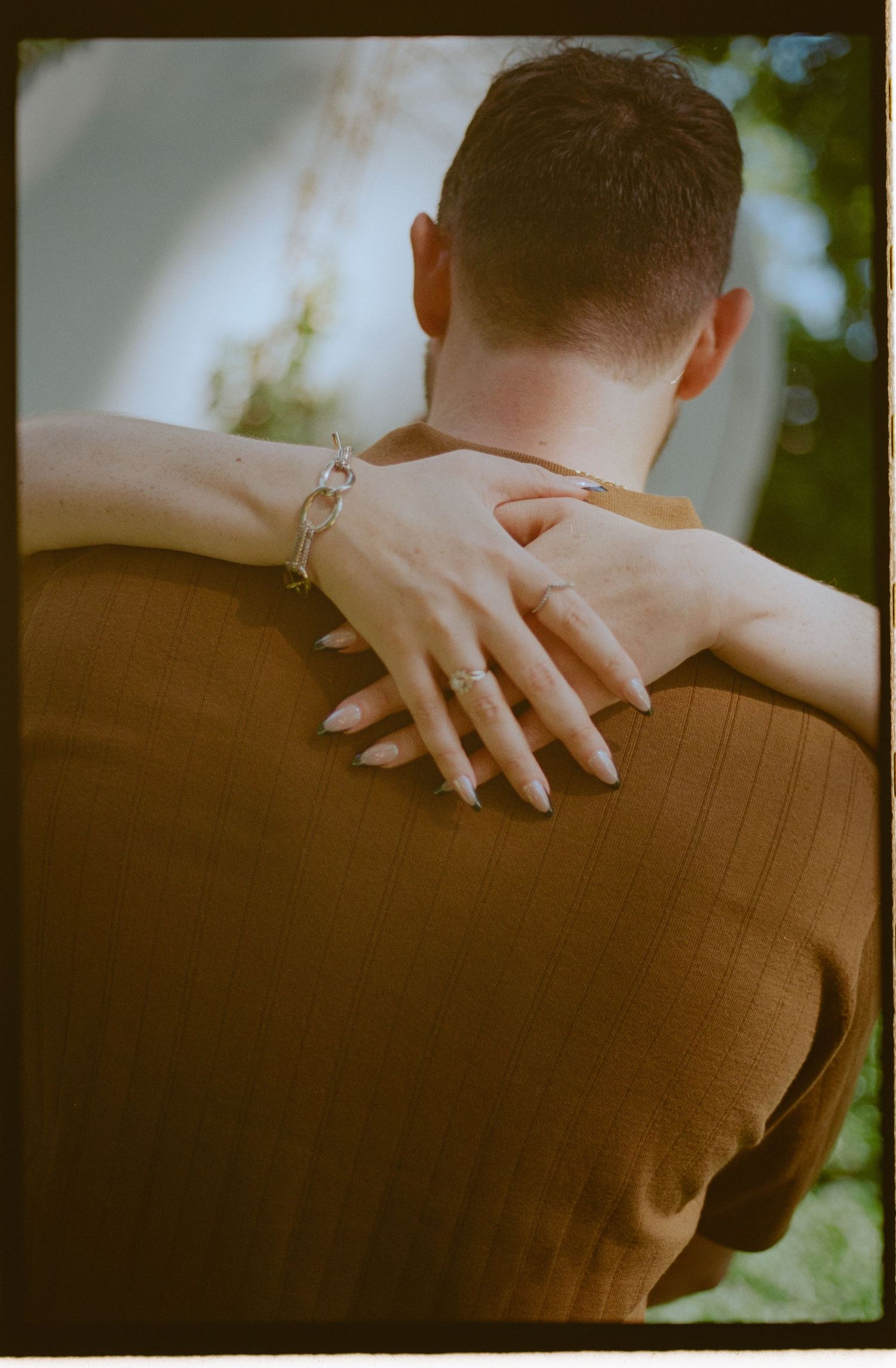 A woman with manicured nails and a silver bracelet embraces a man from behind, her hand resting on his shoulder.