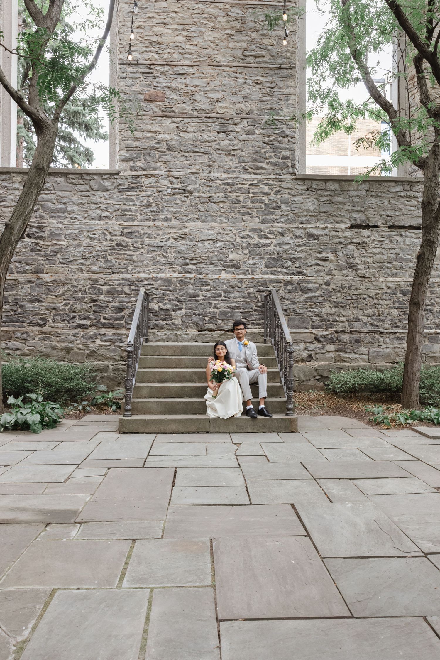 A couple sits on stone steps against a textured stone wall surrounded by trees in an outdoor setting.  St. Josephs park wedding photographer