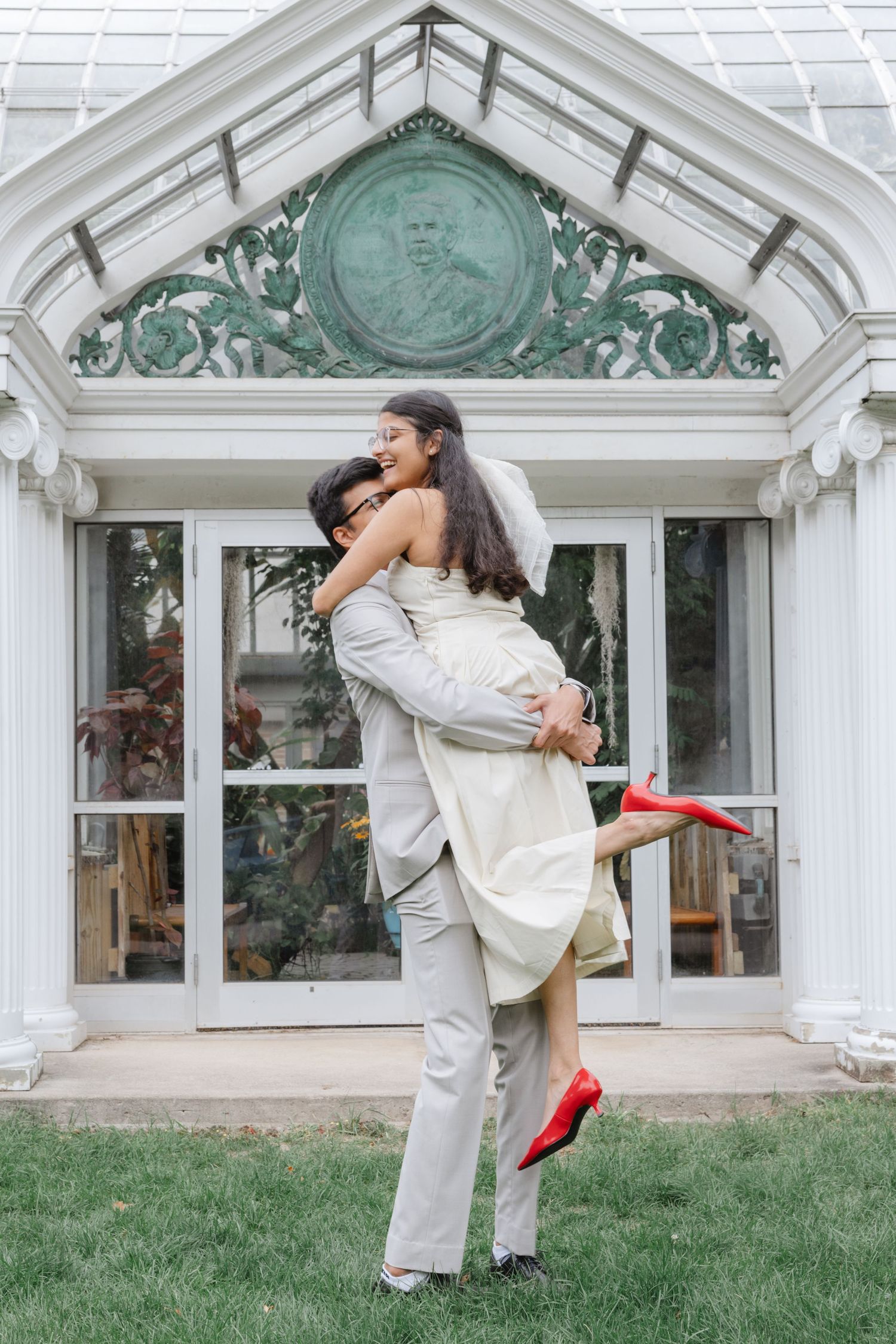 A couple in white attire dips and dances playfully in front of a white building with green architectural details.  Rochester conservatory we