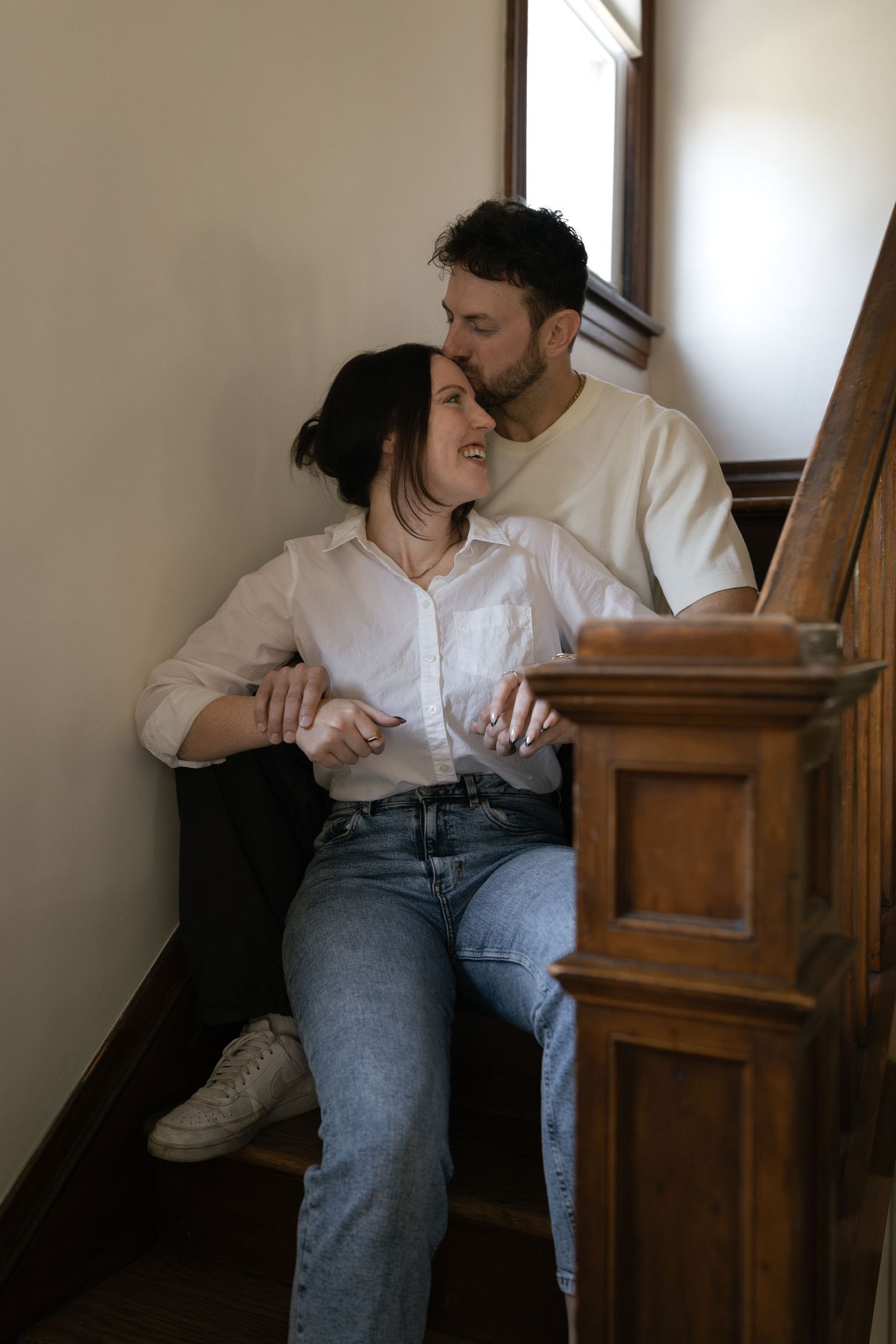 Couple sitting on wooden staircase, man kissing woman on cheek while she smiles, both wearing casual white shirts and jeans.