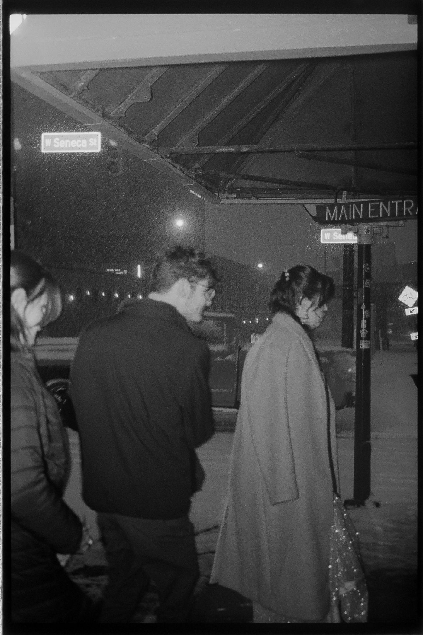 A dimly lit nighttime scene shows people walking near a building entrance with illuminated signage.