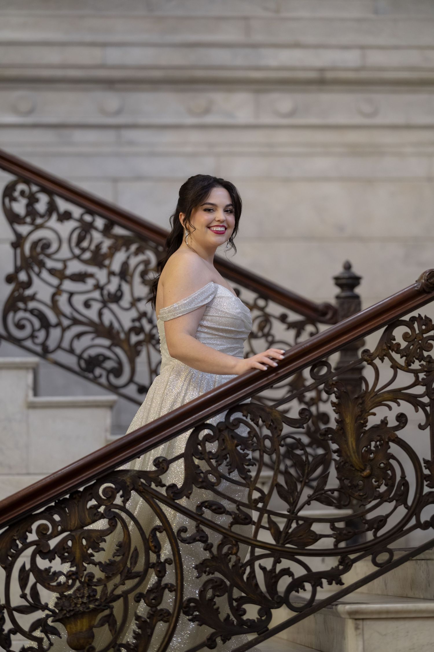 A woman in a white dress stands on an ornate wrought-iron staircase, smiling and looking at the camera.