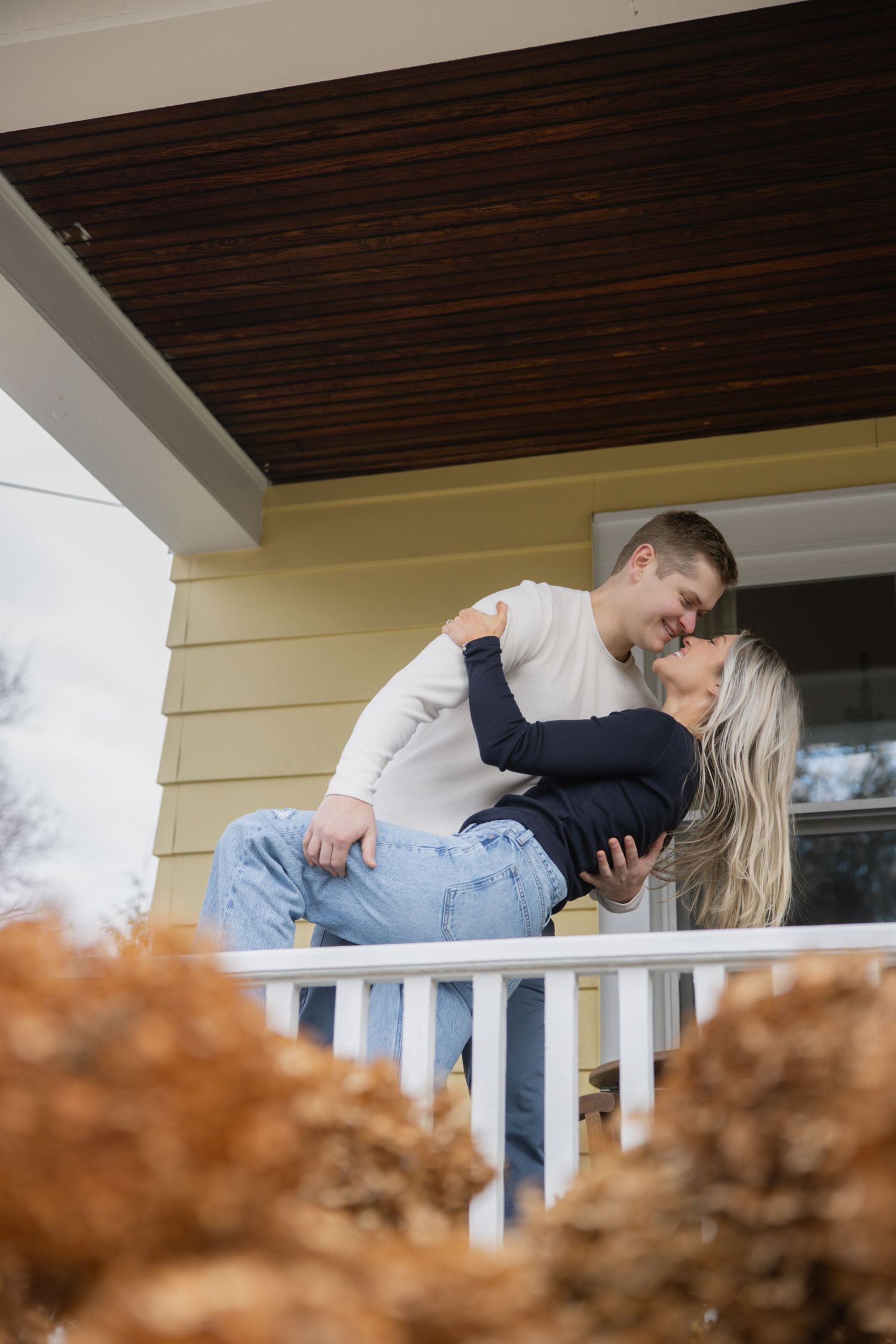 A romantic moment shared on a white porch with autumn foliage in the foreground. Fairport NY home engagement photography