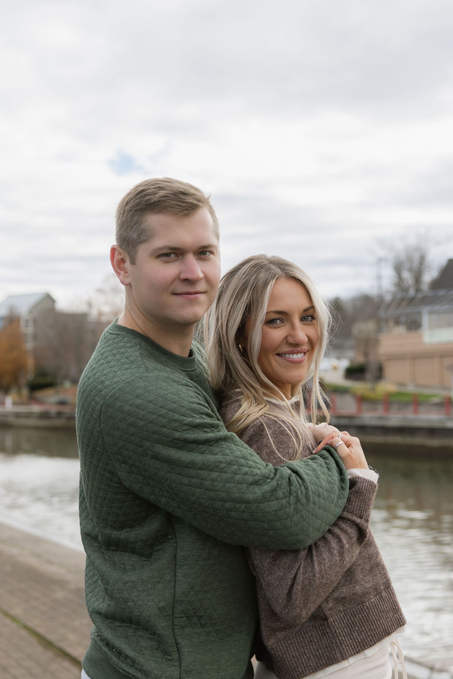 A couple poses for a portrait by the river with cloudy skies and autumn foliage in the background. Film wedding photographer Rochester  NY