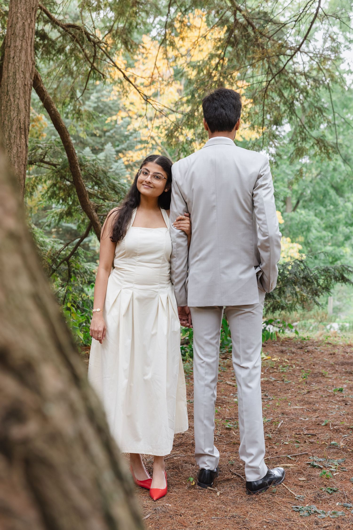 A couple in white and gray attire walking through a forest of tall evergreen trees. Rochester conservatory elopement photographer