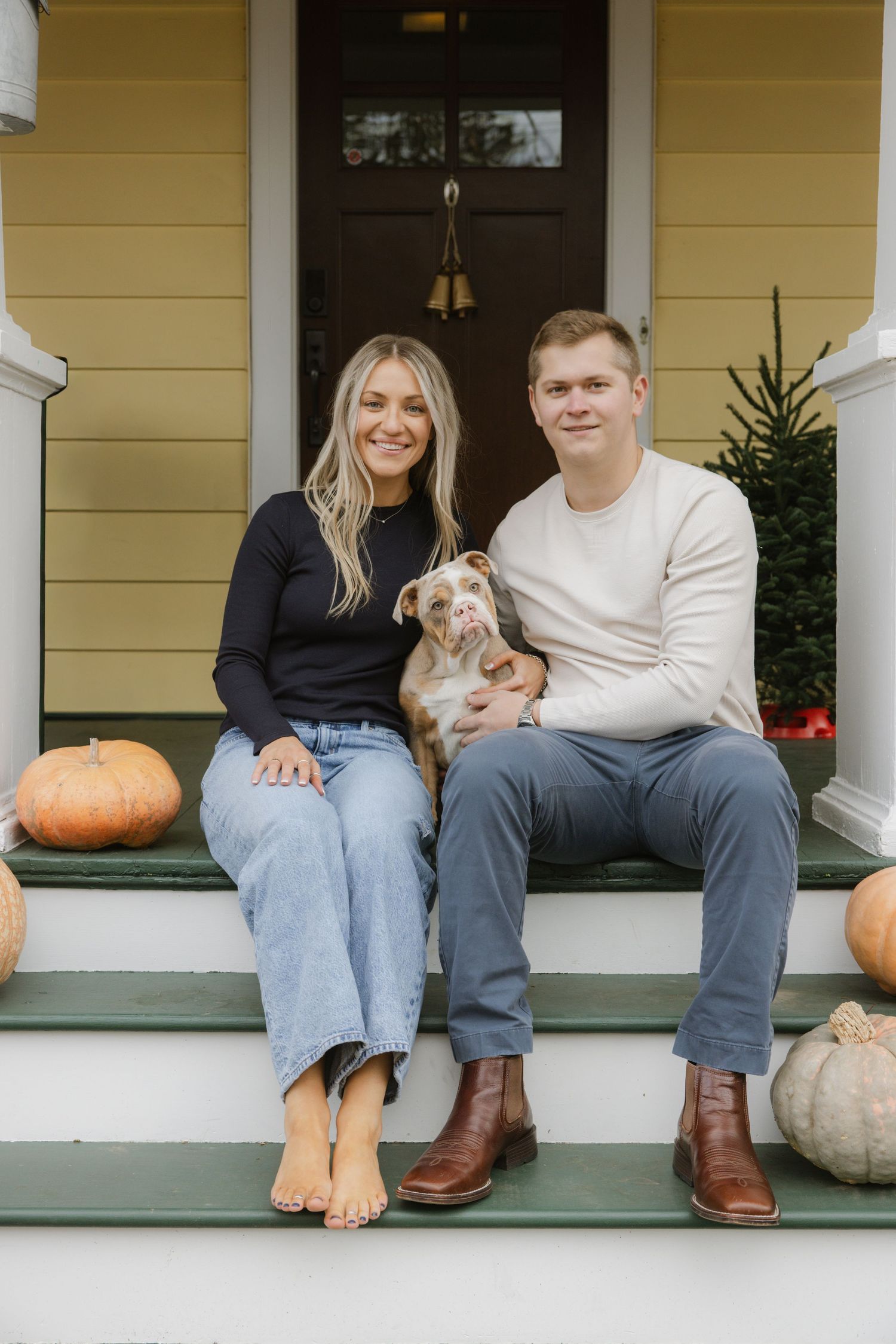 A couple and their dog pose on their porch steps decorated with fall pumpkins. Rochester NY home engagement photography
