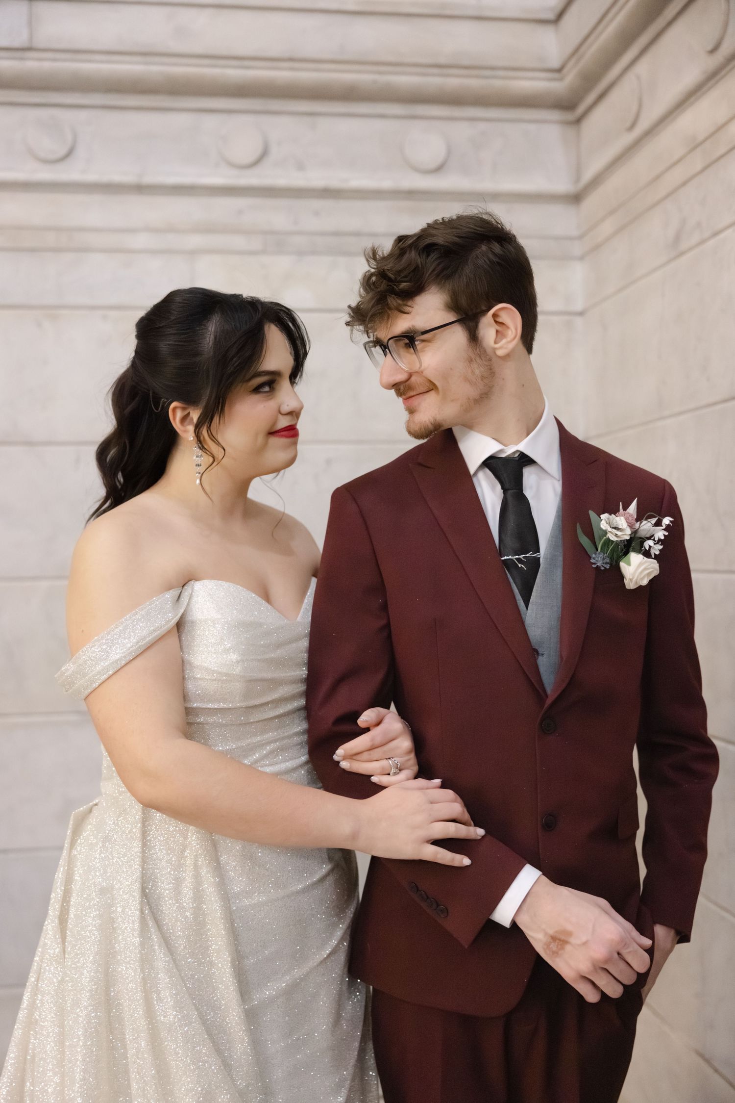 A romantic wedding couple in burgundy suit and white dress share intimate moments against a light colored wall.