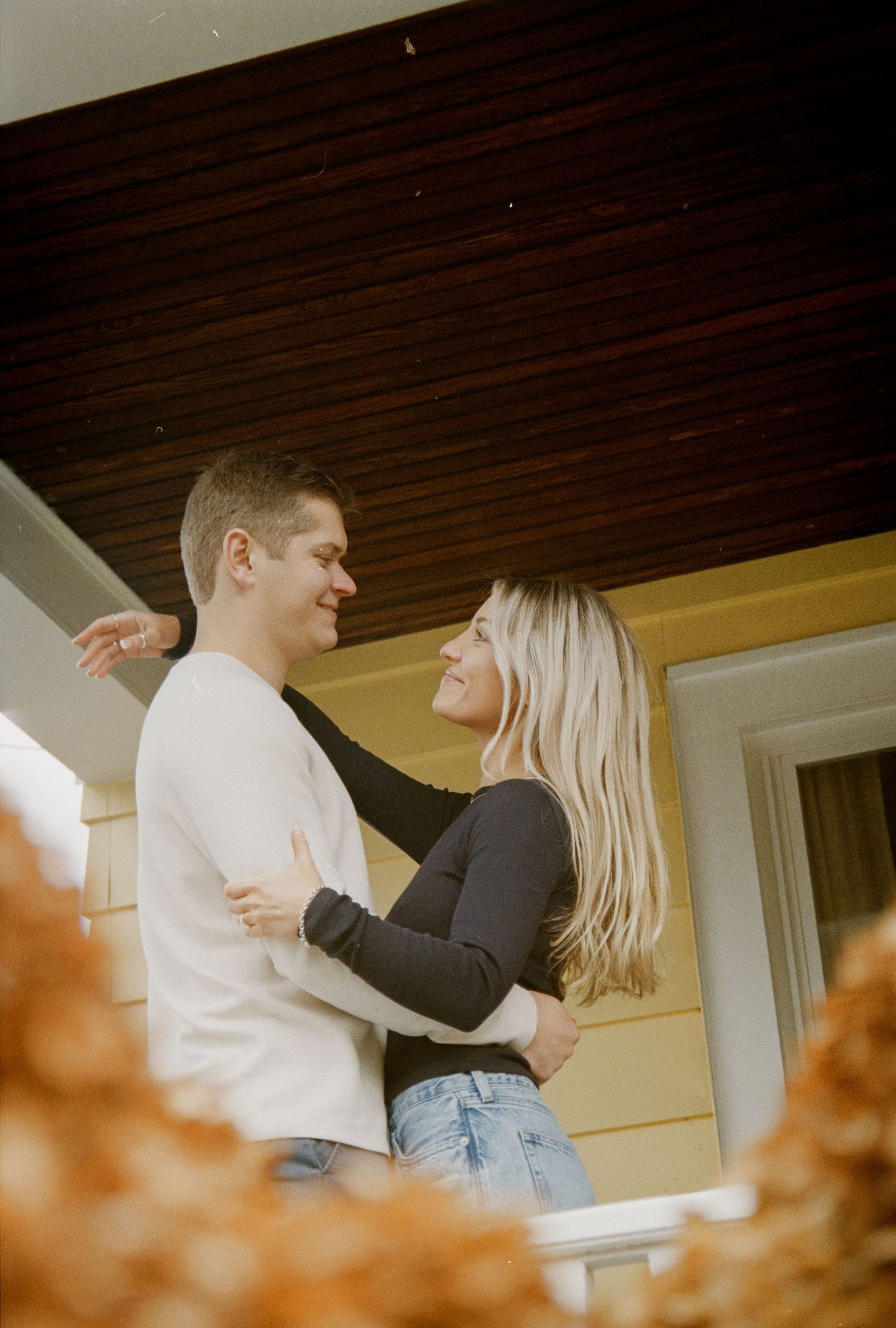 A couple embraces on their front porch in warm autumn lighting. Rochester NY home engagement photography