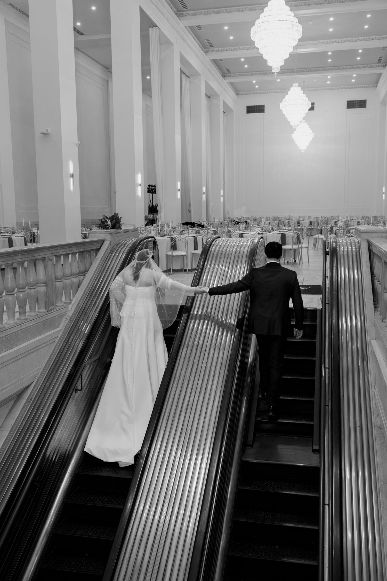Wedding couple poses in a grand white hall with crystal chandeliers and wooden church pews. Arbor Midtown wedding photographer