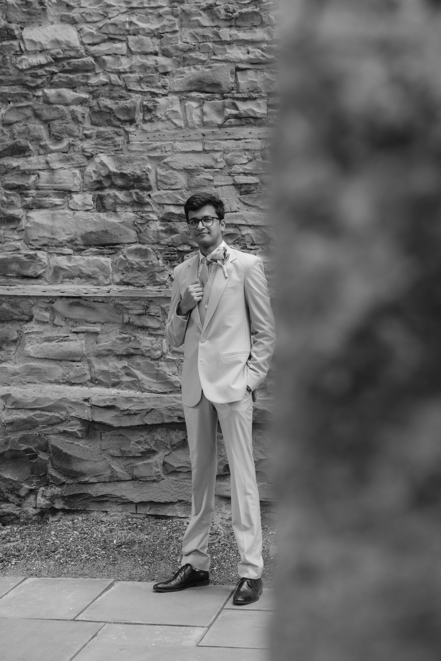 A person in a light colored suit poses against a textured stone wall in black and white.  Fun elopement photography Rochester NY