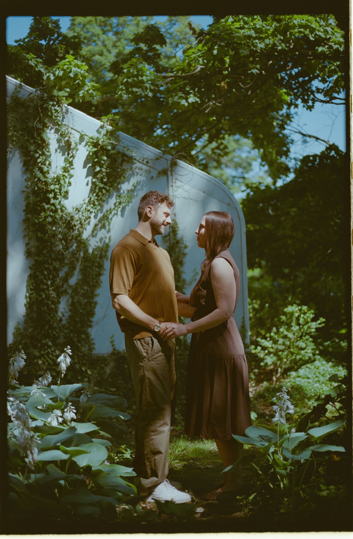 A couple holds hands and gazes at each other in a lush garden with ivy-covered walls and green foliage.