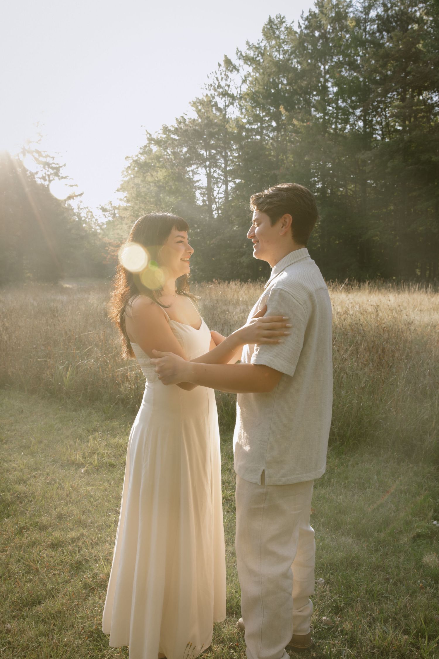 A romantic couple embraces in a sunlit meadow surrounded by tall pine trees at golden hour.  Rochester NY authentic engagement photography