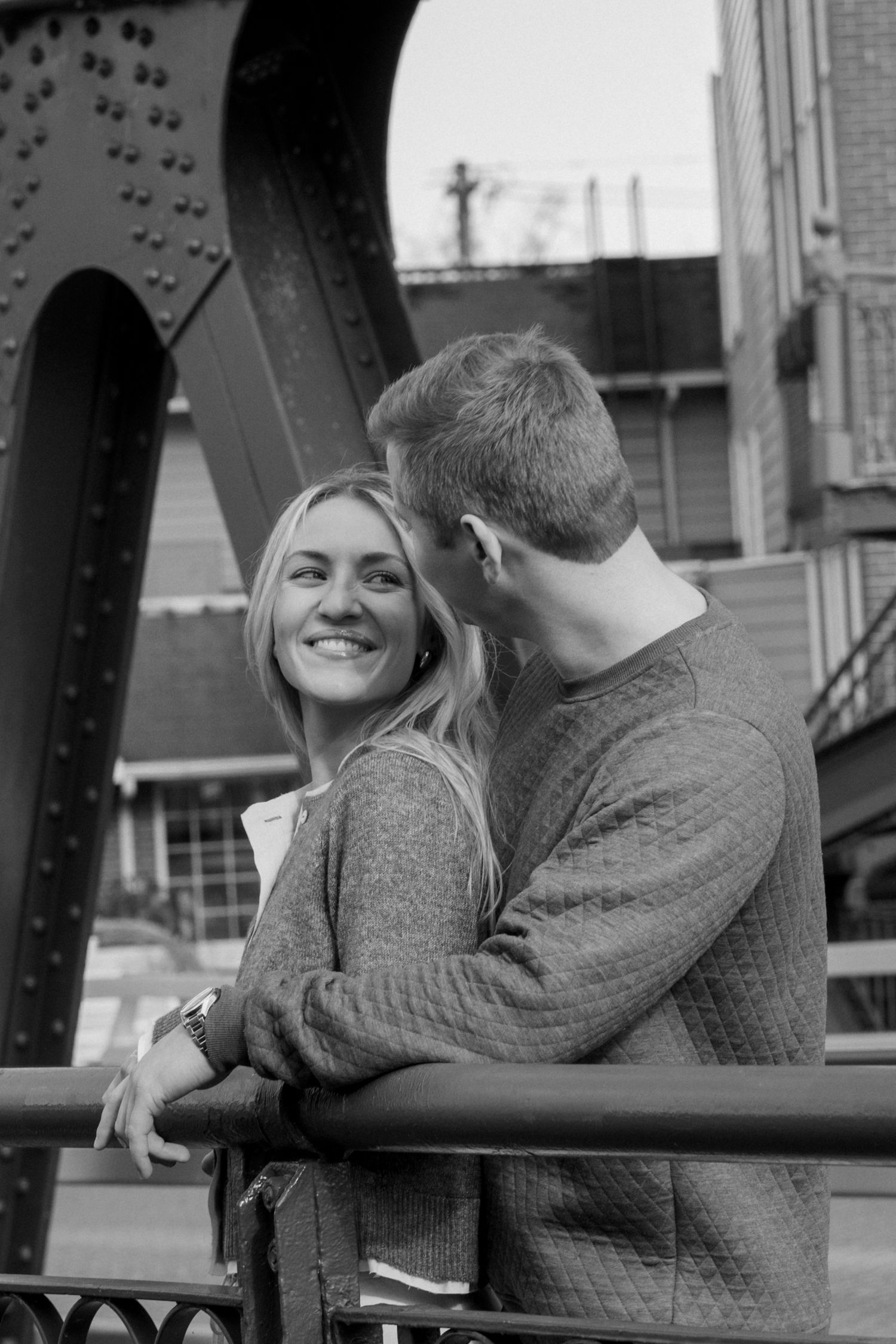 A couple shares an intimate moment against an industrial bridge backdrop in black and white.
