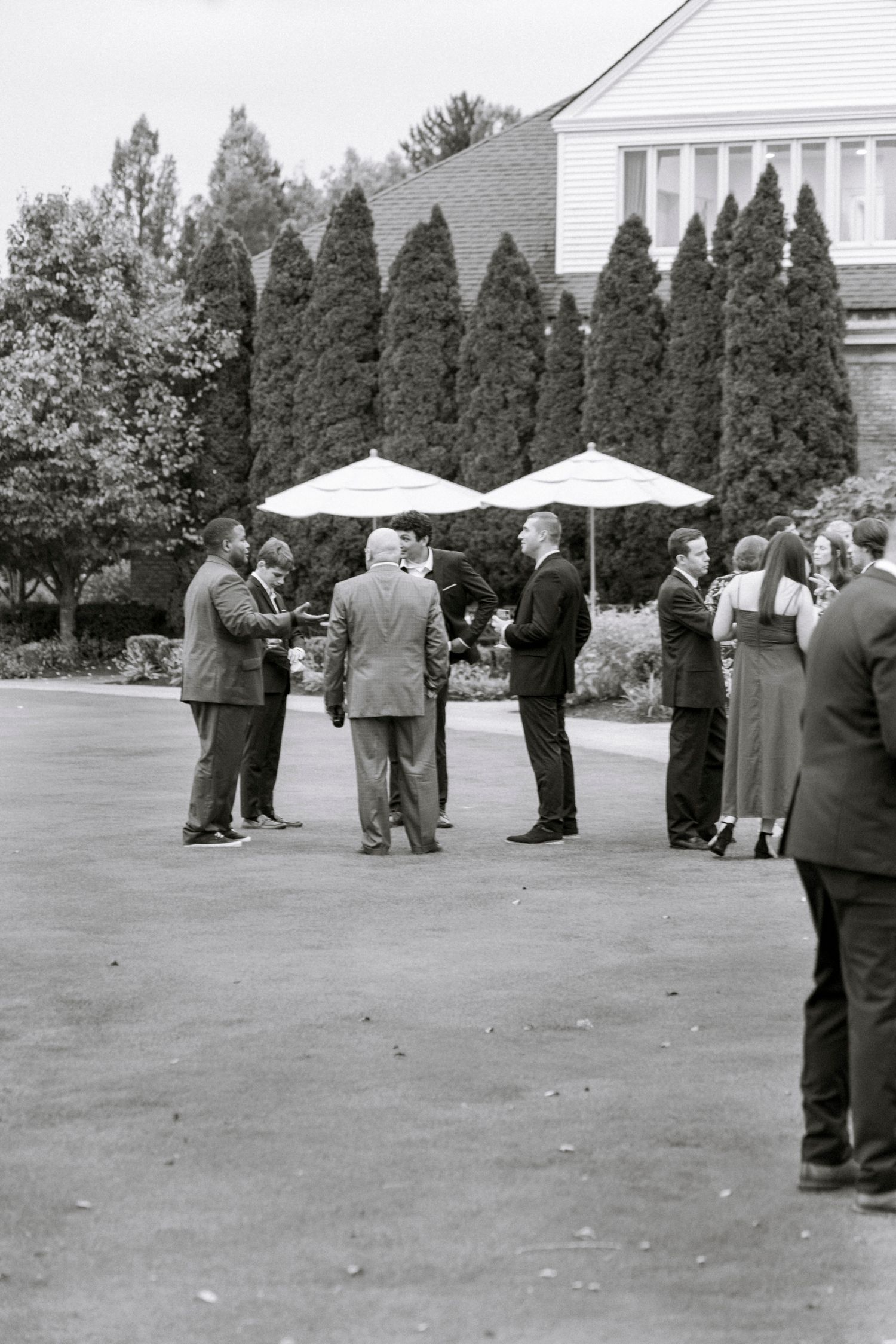 Black and white photo of wedding guests mingling on a lawn near a white building with cypress trees. Rochester NY documentary wedding photog