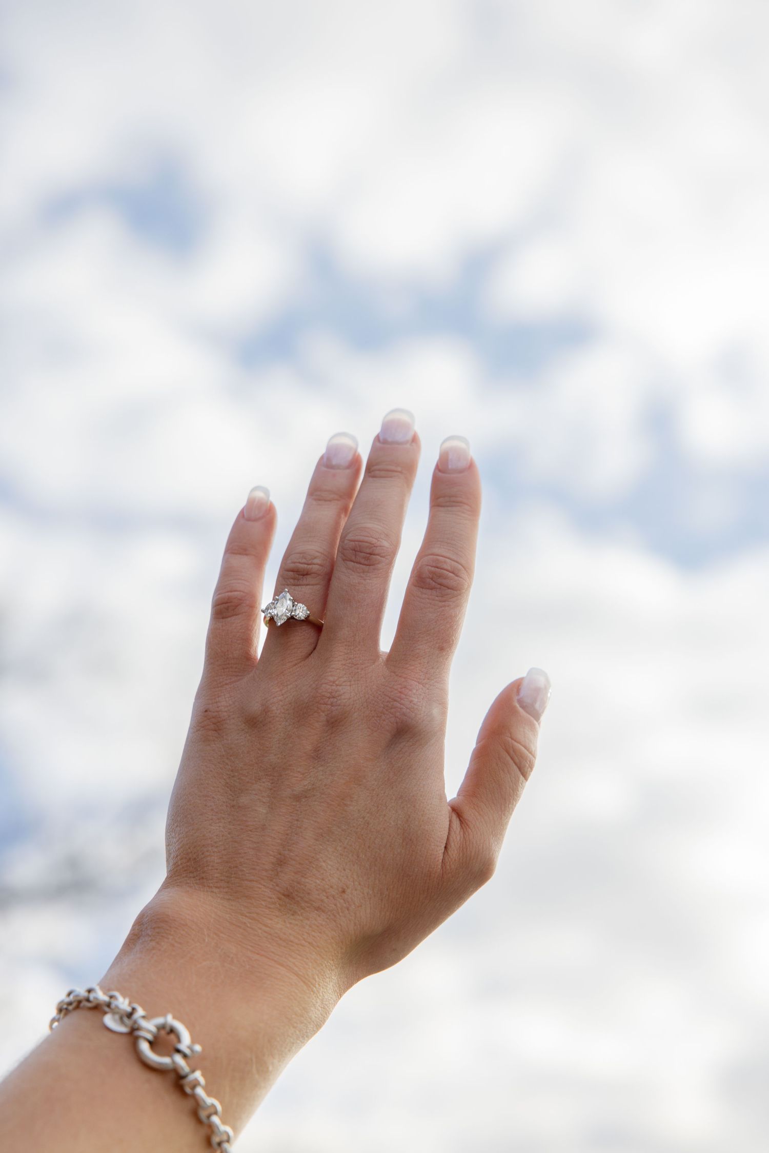 Hand with engagement ring held up against a cloudy sky background.