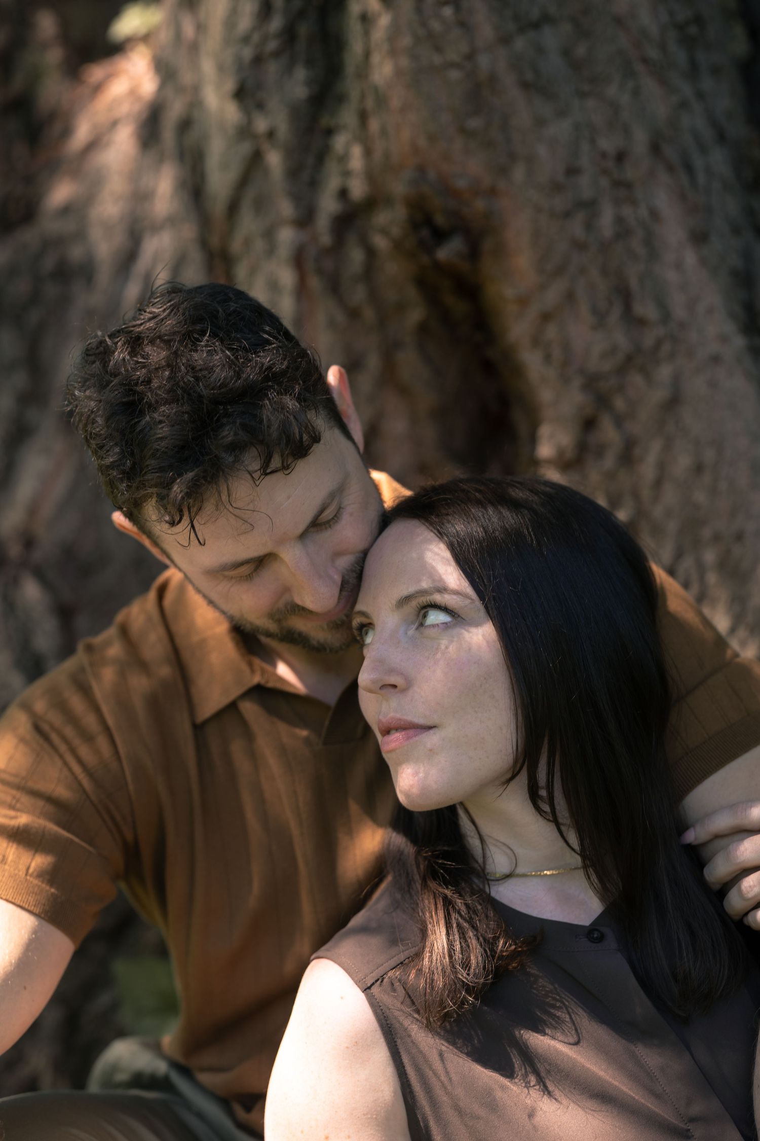 A couple sits intimately near a large tree, the man whispering to a dark-haired woman gazing upward.
