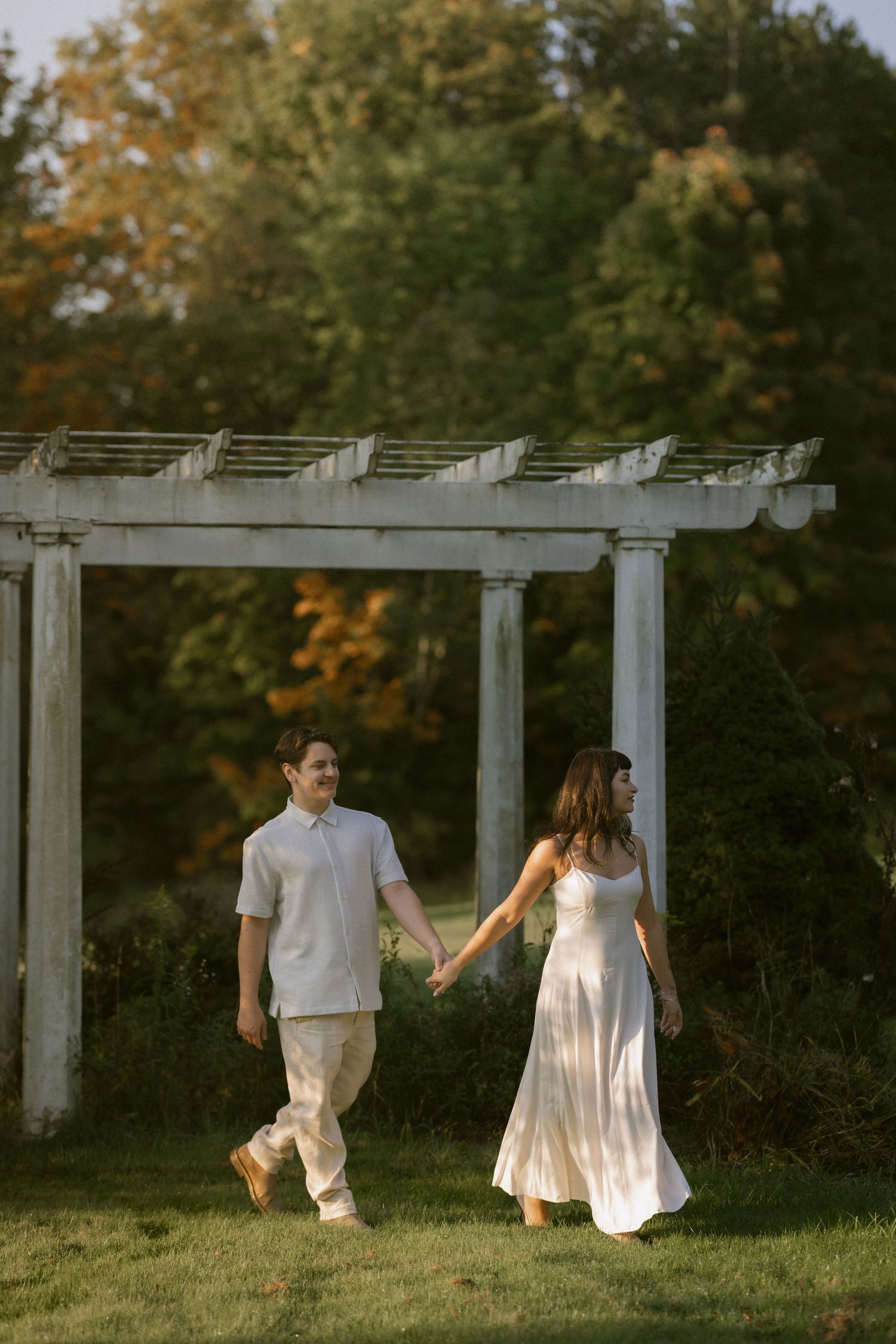 A couple in white outfits walking and holding hands under a white pergola in a garden setting. Rochester NY authentic engagement photography